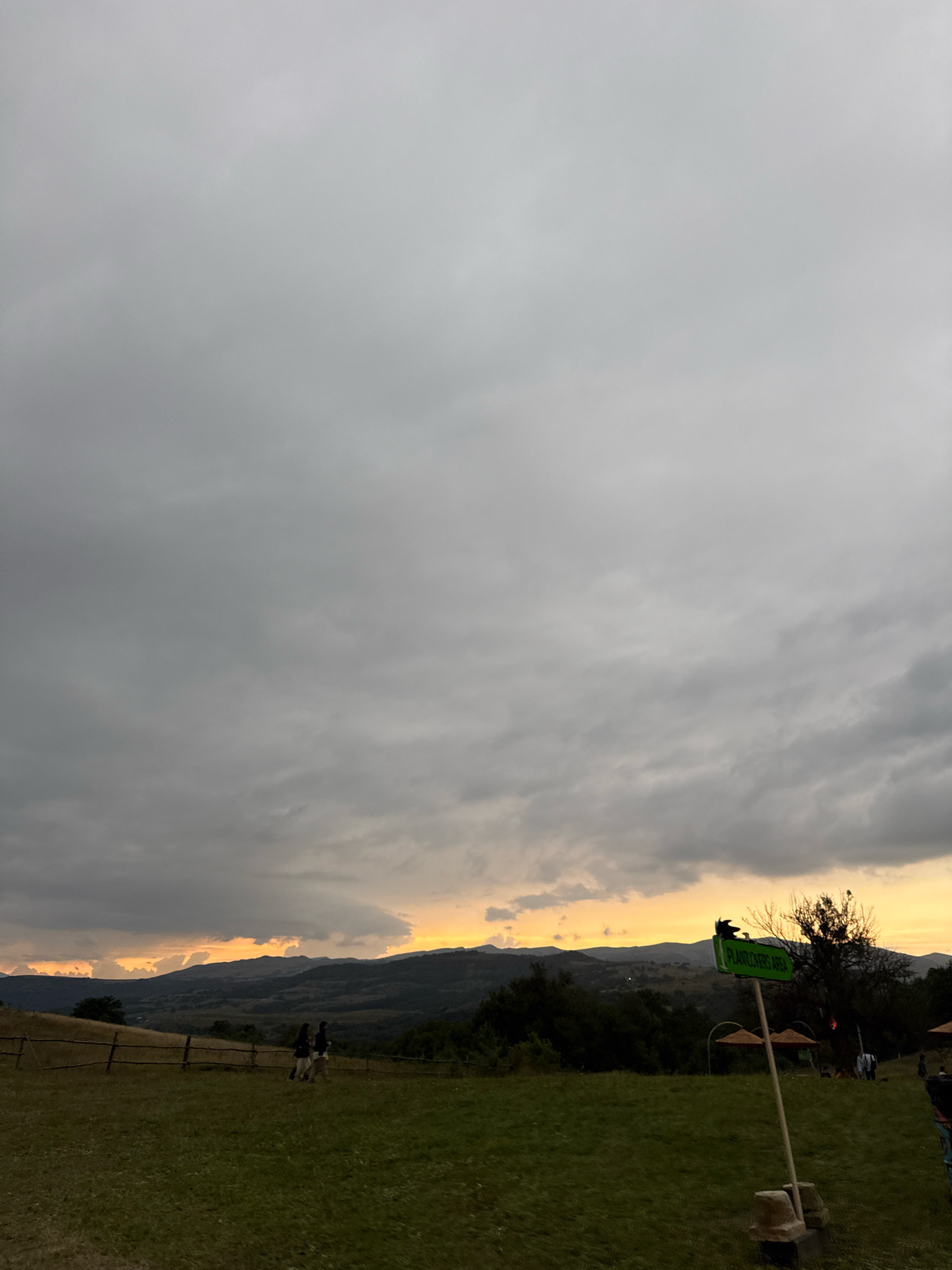 The image depicts a cloudy sky over rolling hills and a grassy field. In the foreground, a wooden fence runs horizontally. Two figures are walking on the grass to the left of the fence. To the right, a green sign with white text reads "PLANT LOVERS AREA" and is attached to a pole. In the middle ground, several parasols are set up on the grass. The background consists of a mountainous landscape under a dramatic sky, with hints of orange and yellow from a setting sun. The location is identified as Orbeti, Georgia.