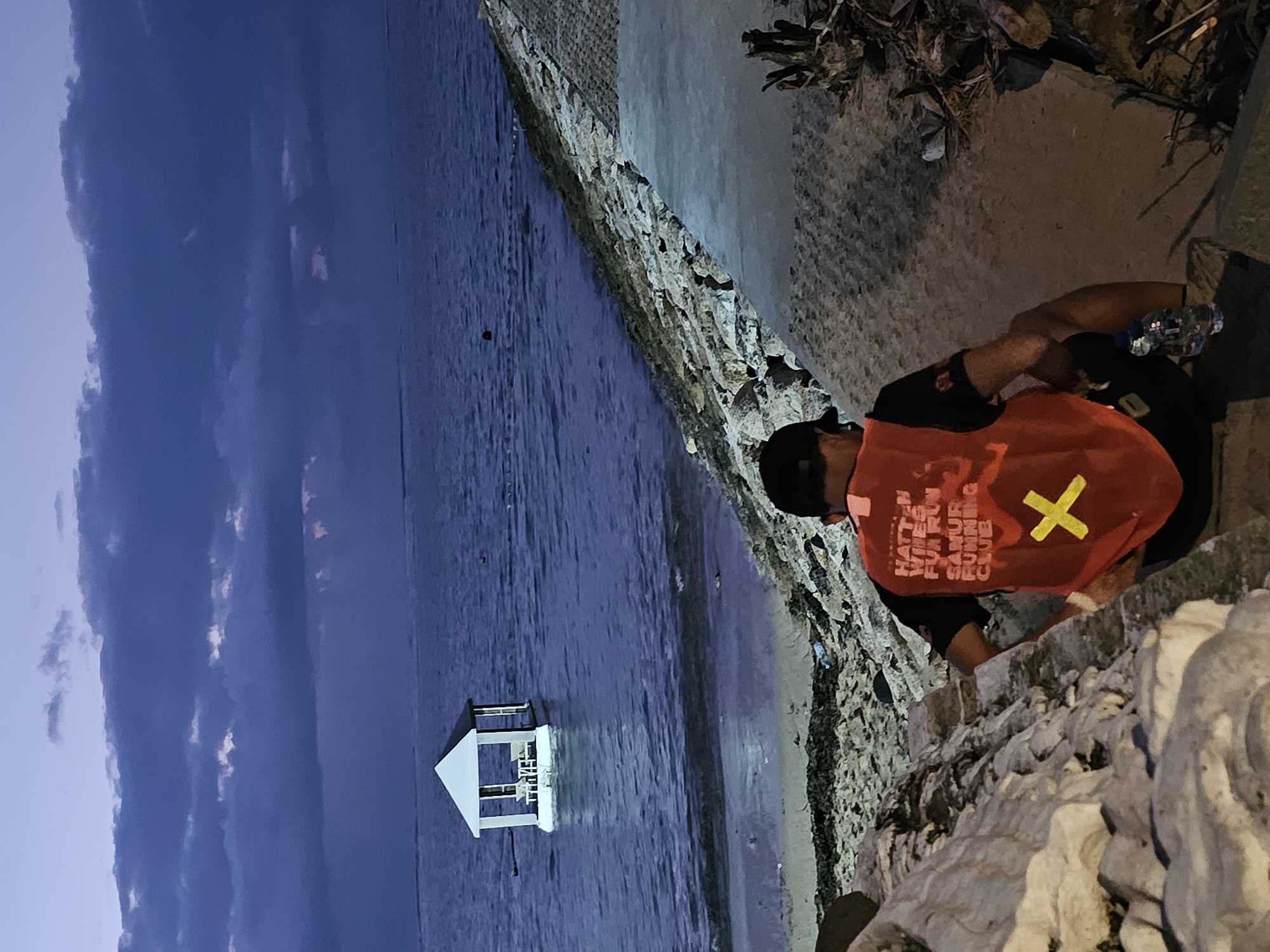 The image depicts a dusk scene at a waterfront location in Denpasar, Indonesia. In the foreground, a person wearing an orange vest with lettering and a yellow cross is seated on a concrete ledge, facing away from the camera. The vest appears to be part of a uniform or event attire. A plastic water bottle is visible next to the person. The environment includes a sandy beach area to the left, transitioning to a rocky embankment and a paved walkway on the right. In the middle ground, the ocean stretches out, calm with gentle ripples. A small, white gazebo with a dark roof is situated on a floating platform in the water. The background features a twilight sky with dramatic, dark clouds and lighter patches where the sun may have recently set or is about to rise. The overall atmosphere is serene and slightly moody due to the low light conditions.