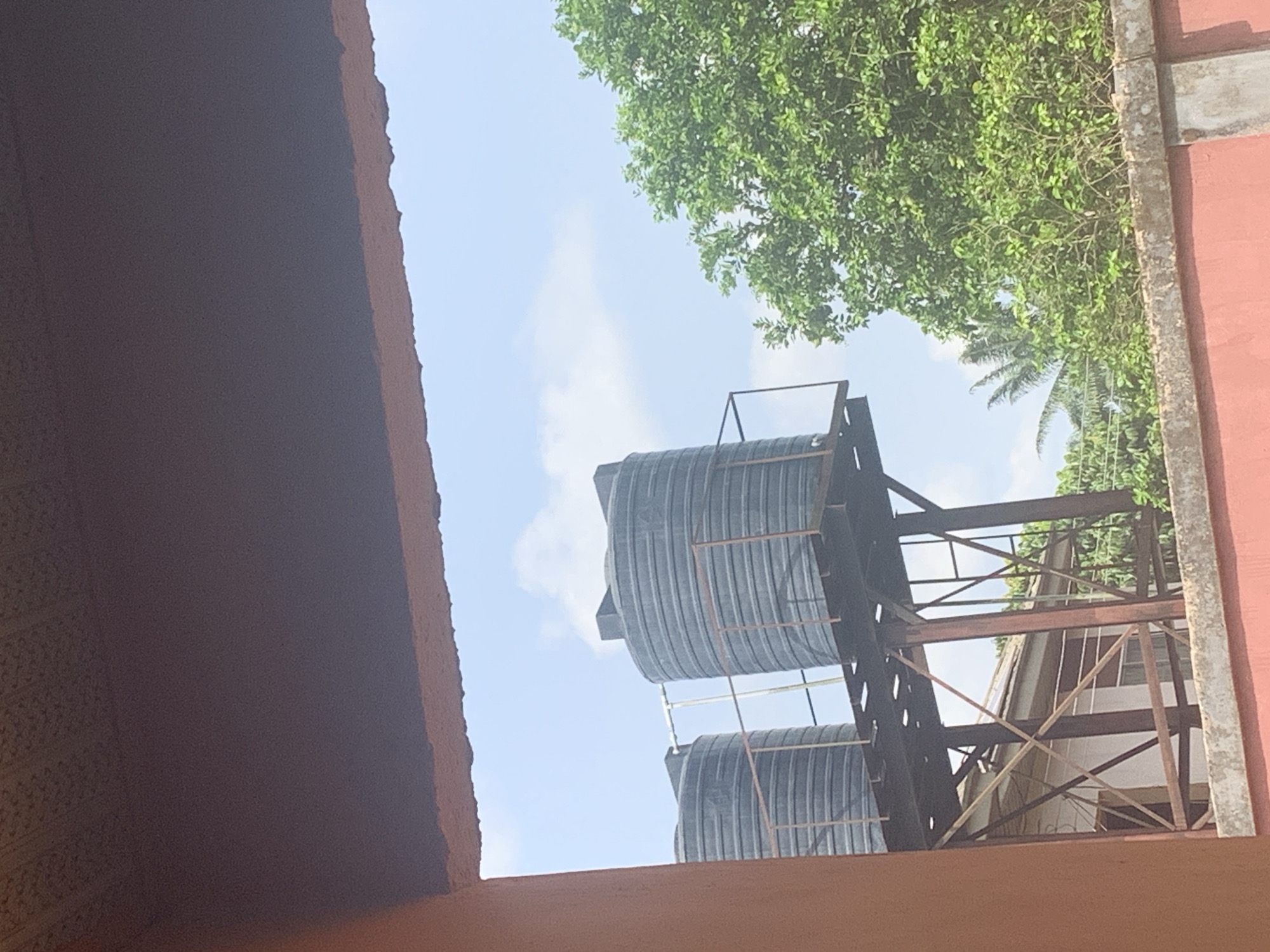The scene depicts two black, cylindrical water tanks stacked atop a rusty, metal frame structure. The view is partially framed by a dark-brown structure on the left and a red wall with a tree growing behind it on the right. The sky is visible between the structures, featuring light clouds. The environment appears to be in Umu-Neke, Nigeria, given the prompt's directive. The scene lacks any indication of people or any specific interactions.