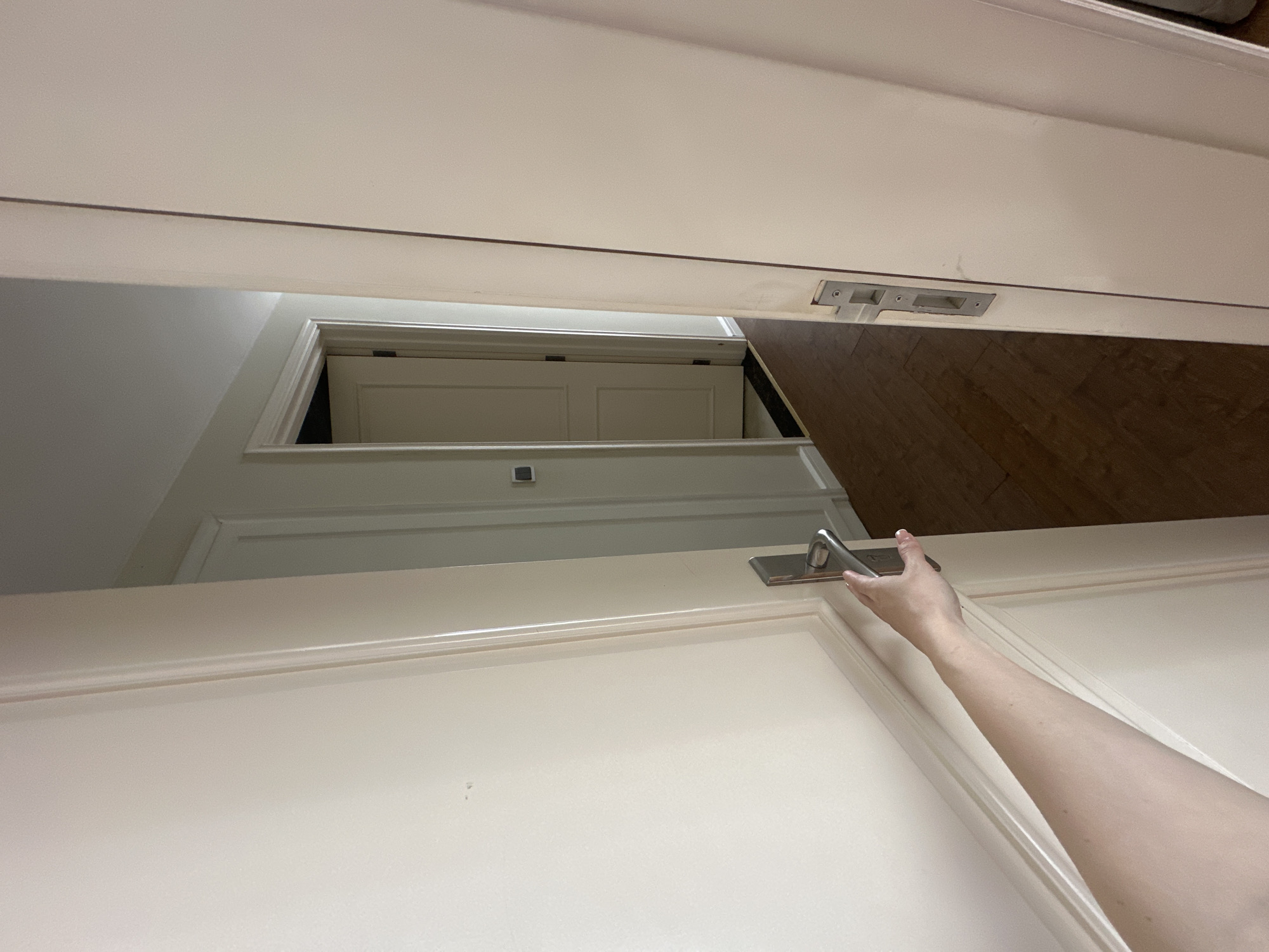 The image presents an unusual, high-angle perspective looking down into an interior space. In the immediate foreground, a light-colored, possibly white or off-white, paneled door is partially open, viewed from its top edge. A modern, silver-toned door handle and its latch mechanism are prominently featured on this door. A person's arm, with fair skin, extends into the frame from the right, with fingers gripping or resting on the handle, indicating the act of opening or closing the door.
Through the gap created by the open foreground door, another light-colored paneled door is visible further down and to the left, framed by a doorway, suggesting another room or passage beyond. This background door appears to be closed or partially closed. To the right and below the foreground door, a section of warm-toned wooden flooring is visible. The surrounding walls are also light-colored. The overall scene suggests a contemporary indoor setting, possibly a hallway or a room with multiple entryways. The unusual vantage point creates a striking sense of depth and architectural complexity. No text is visible, and the time of day is not discernible due to indoor lighting.