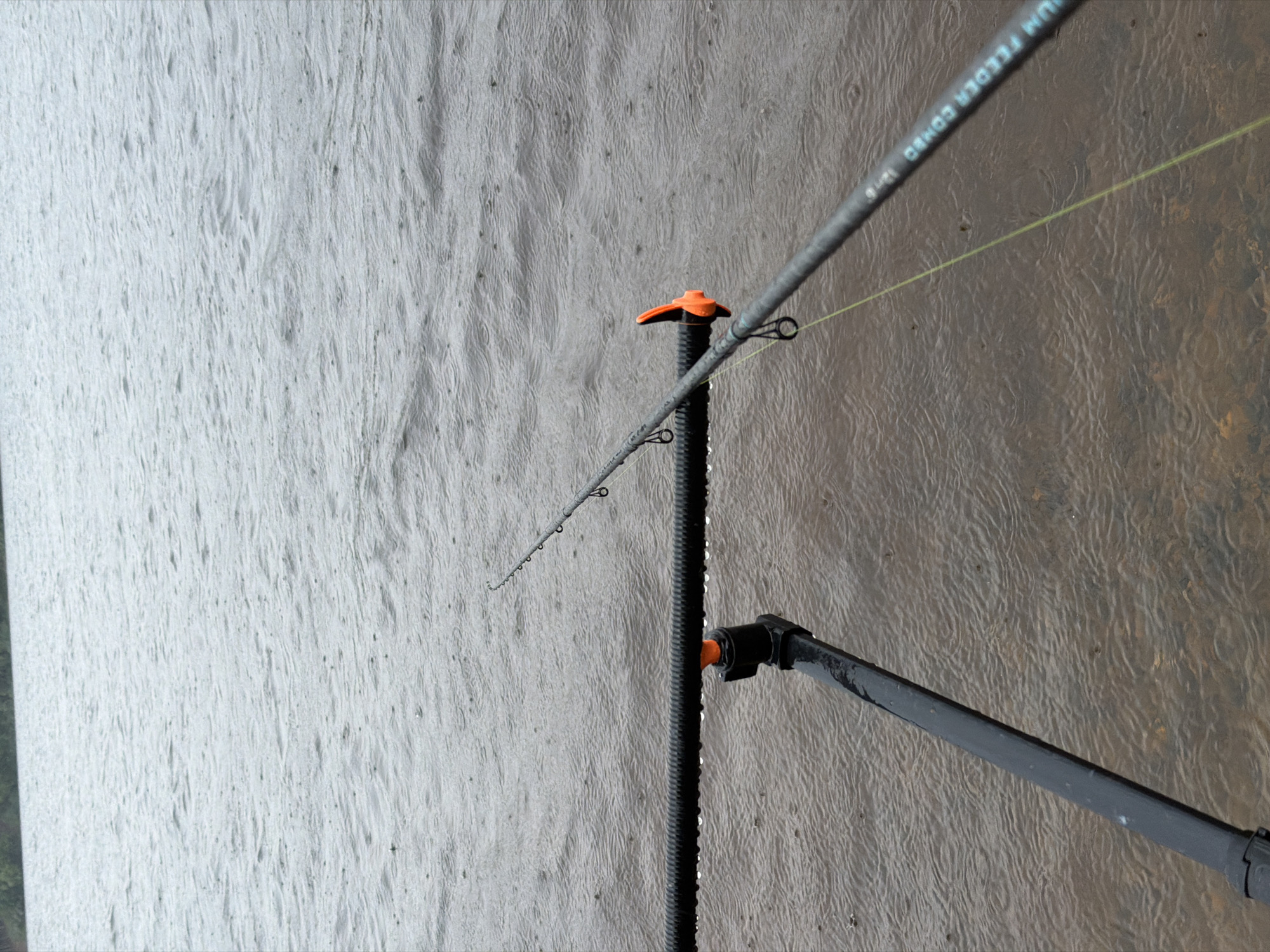 The image depicts a fishing rod resting on a rod pod, angled over a body of water. The water's surface is disturbed by raindrops, indicating it is raining. The background shows a blurry line of trees or land. The rod pod is black and made of metal, with a ribbed surface on the horizontal bar. An orange component is visible on the rod pod. The fishing rod is grey with black rings. A yellow fishing line extends from the rod into the water. The scene appears to be a bank of a river or lake, possibly in an outdoor recreational area. The location is identified as Chaikovsky, Russia.