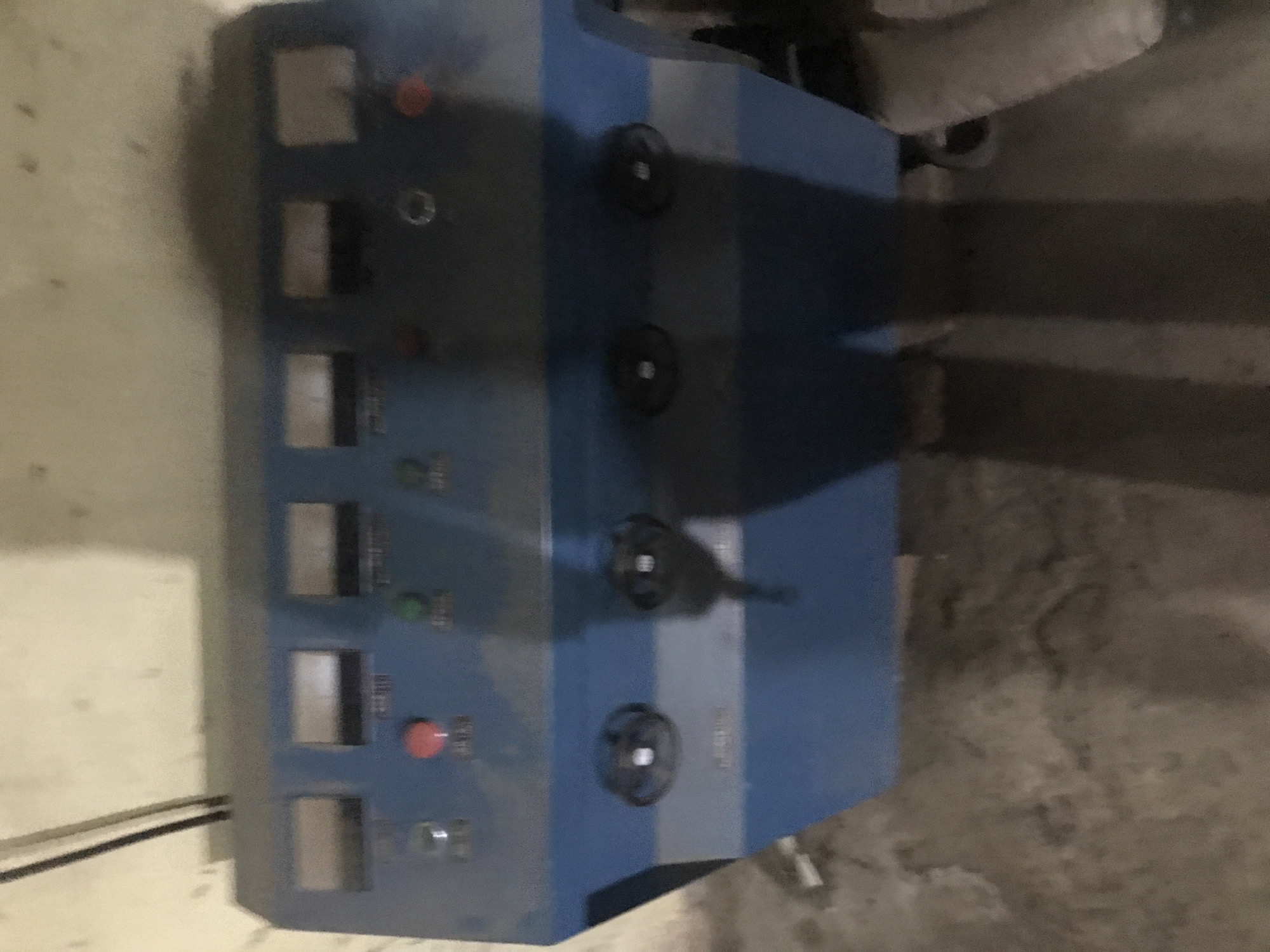 A blue control panel, possibly part of an industrial or scientific apparatus, is situated against a light-colored, textured wall. The panel features multiple displays, buttons, and what appear to be knobs or dials. The surface of the panel is dusty, indicating it is not in active use or is in an environment with significant dust accumulation. The floor in front of the panel is rough, concrete or dirt, with some debris present. Shadows are cast on the floor, suggesting lighting from above and to the side. The overall impression is of an indoor, possibly disused, industrial or utility space. No specific location in Kabong, Nigeria, can be determined from the image itself.