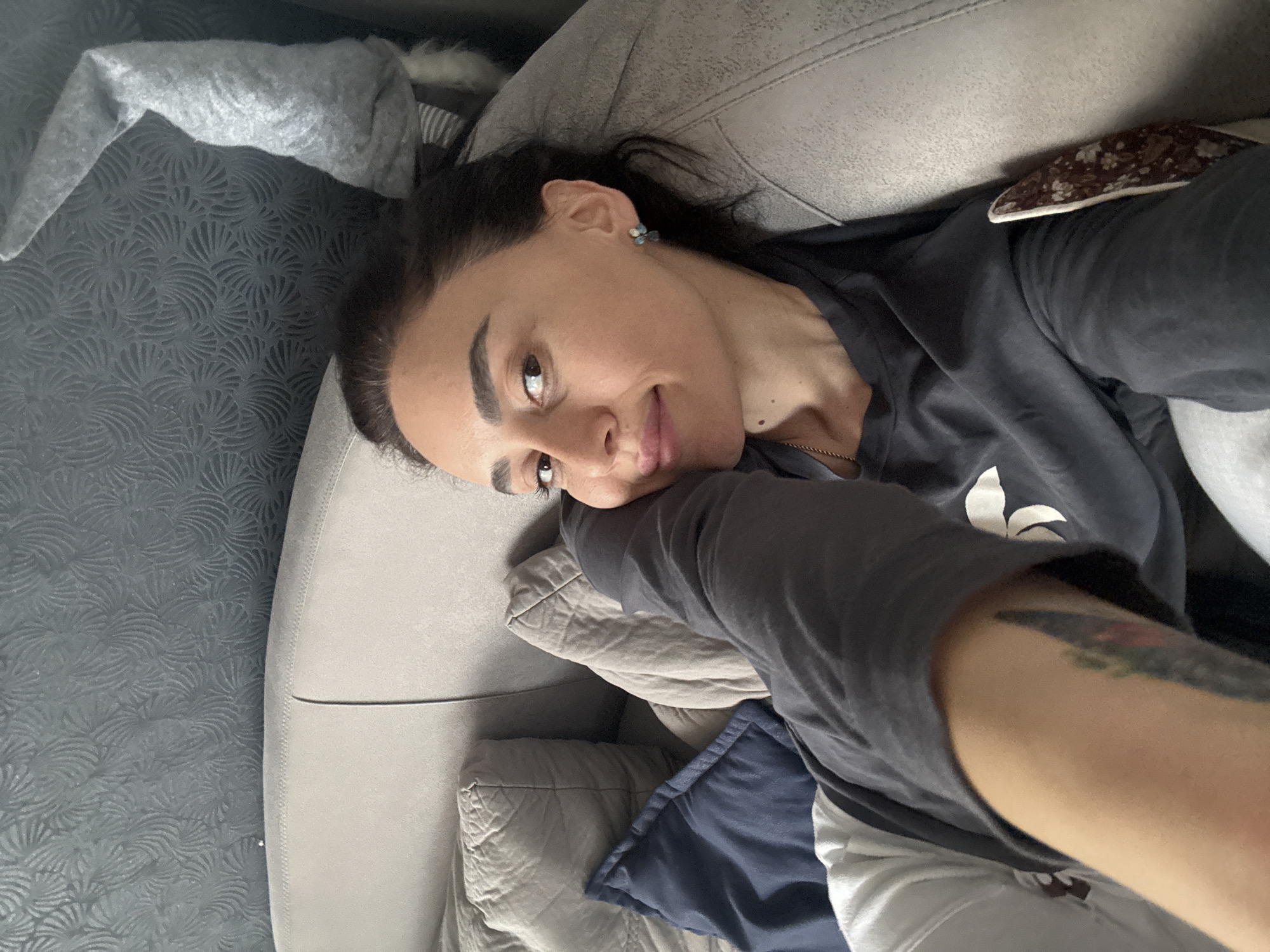 A woman with dark hair and fair skin is lying on a light-colored sofa. She is wearing a dark gray t-shirt and has a tattoo on her left arm. The sofa is placed against a dark gray patterned wall. Pillows are visible on the sofa. The location cannot be confirmed as Tbilisi, Georgia from visual details.
