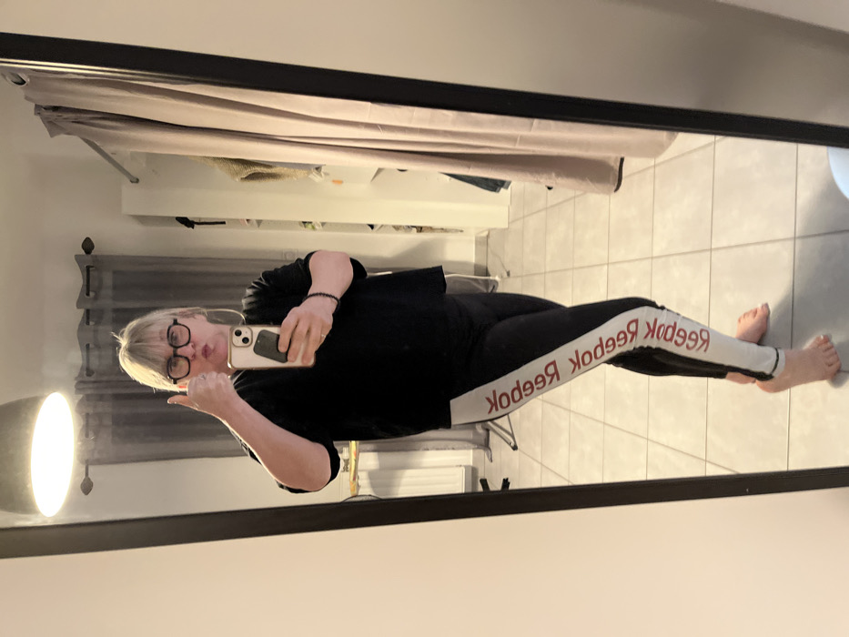 The image is a mirror selfie taken indoors, appearing rotated 90 degrees counter-clockwise.
A person, likely a woman, with light-colored, short-to-medium length hair and dark-rimmed glasses, is visible in the reflection. She is holding a smartphone in her right hand, which is covering part of her face, and her left hand is raised, showing a "thumbs up" gesture. Her lips are slightly pursed.
She is dressed in a casual outfit consisting of a black long-sleeved top and black leggings. The leggings feature a distinctive design: a wide white stripe runs down the outer side of each leg from hip to ankle. Printed repeatedly in red on this white stripe is the word "Reebok." Due to the mirror reflection, the text appears backward as "qooddeЯ qooddeЯ qooddeЯ". She is barefoot, with her feet clearly visible on the floor.
The setting appears to be a room with light-colored, possibly off-white or light grey, tiled flooring and light-colored walls. In the background, light-toned curtains are drawn across a window or opening. To the left of the person (when the image is viewed upright), a large, modern floor lamp with a prominent, circular light source is casting a bright glow. The overall lighting suggests an indoor, possibly evening, setting with artificial light sources. The mirror itself has a thick, dark frame.