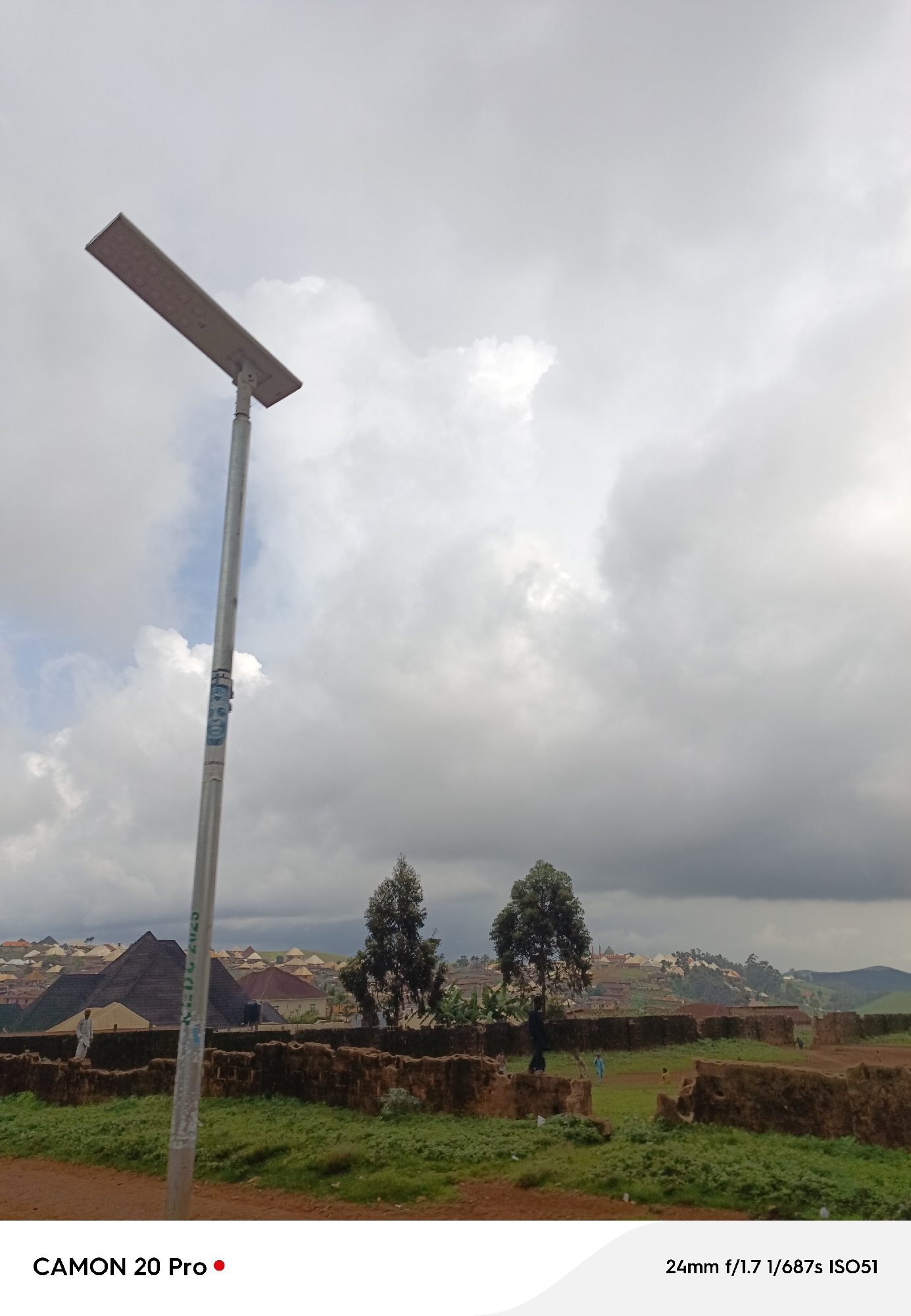 This is a medium shot of a solar-powered streetlamp positioned on the left side of the frame, set against a cloudy sky. The lamp is silver and has a rectangular light fixture. A sticker with a portrait is affixed to the pole. In the background, a settlement with buildings of varying colors and roof types is visible on a hillside. Several trees dot the landscape. In the foreground, there is a low, crumbling brick wall and overgrown green vegetation. A dirt path is in the lower foreground. The image also includes text overlays: "CAMON 20 Pro" in the lower left corner and "24mm f/1.7 1/687s ISO51" in the lower right corner. The location is specified as Nguroje, Nigeria.