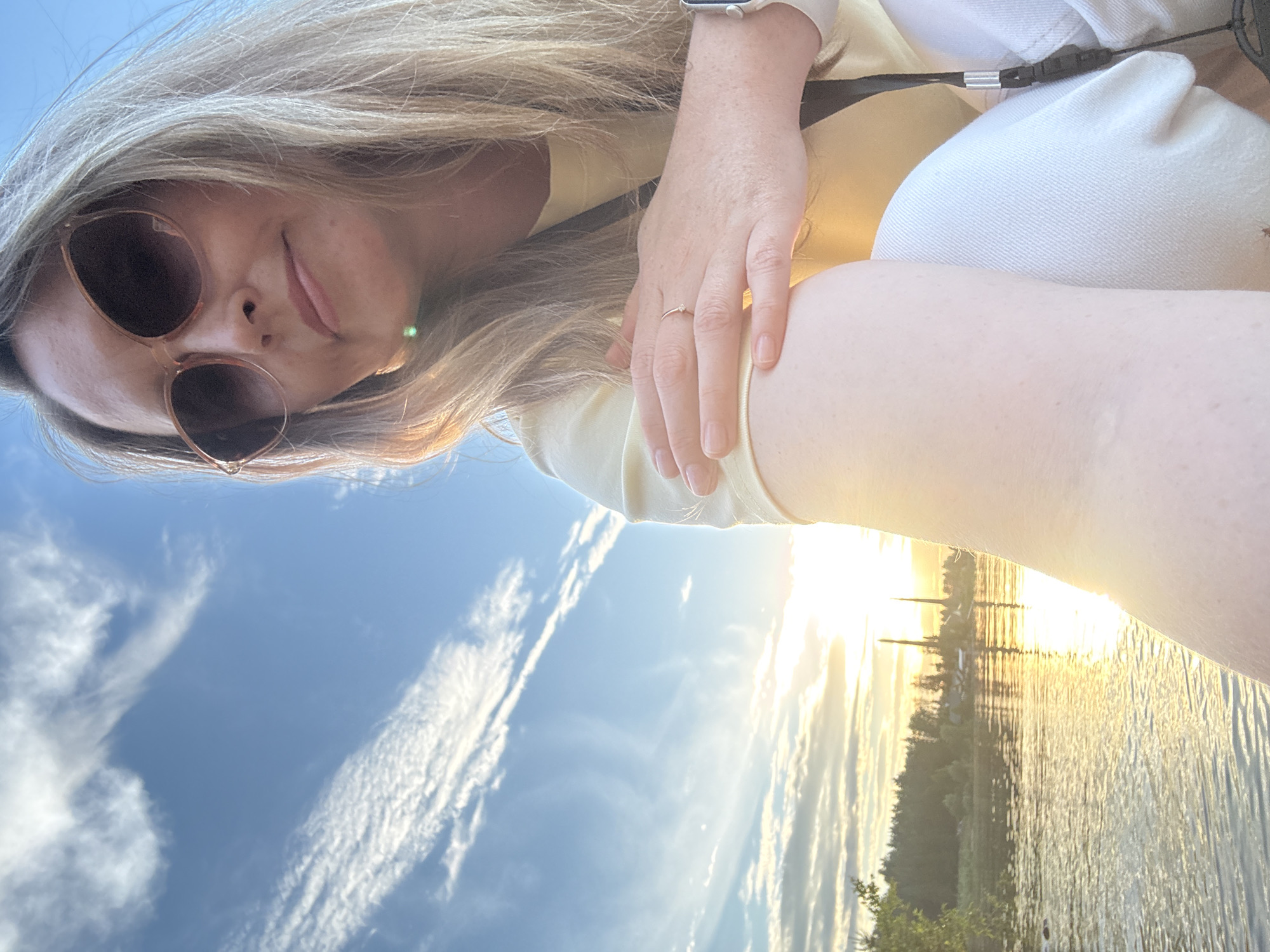 A woman is taking a selfie outdoors, with a bright sun setting behind her. She is wearing sunglasses and a yellow shirt, and has blonde hair. The background shows a body of water with trees on the far shore. The sky is blue with wispy clouds. The sun is low on the horizon, casting a warm glow on the scene. The woman appears to be relaxed and happy. The location is likely near a lake or river. The time of day is sunset.