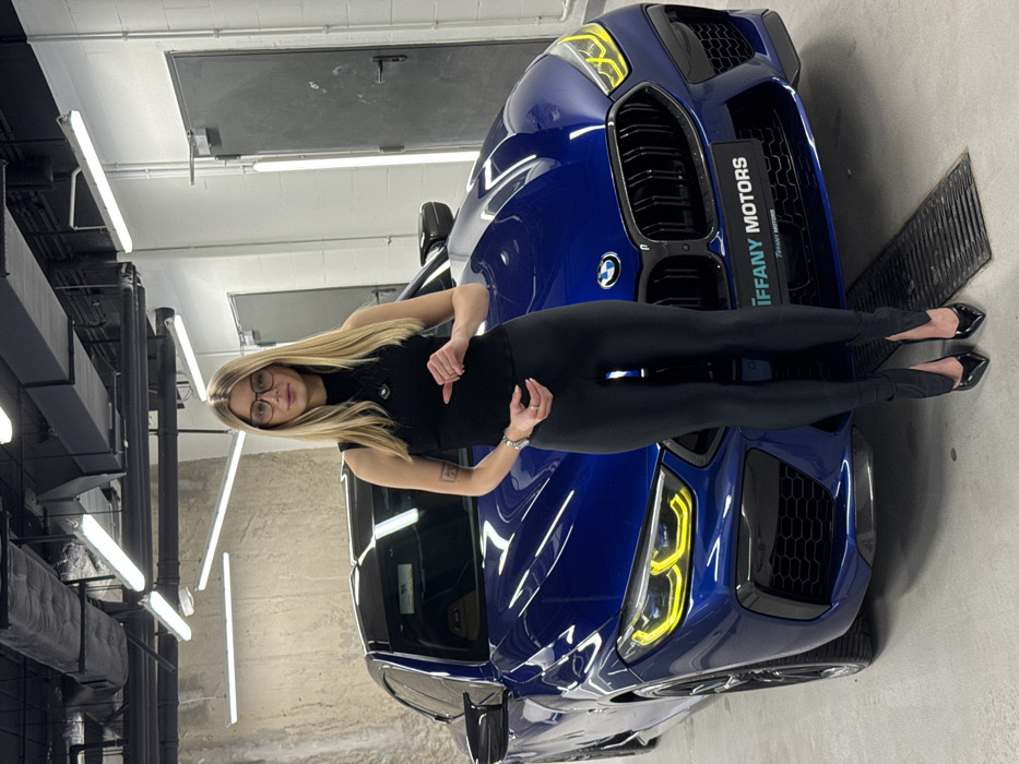 A blonde woman in black leggings and a black top stands in front of a blue BMW with the "Iffany Motors" logo. She wears glasses and points downward with her left hand. The car is parked in a garage-like space with fluorescent lighting and a grate near the front of the car. The image appears to be rotated 90 degrees counter-clockwise.