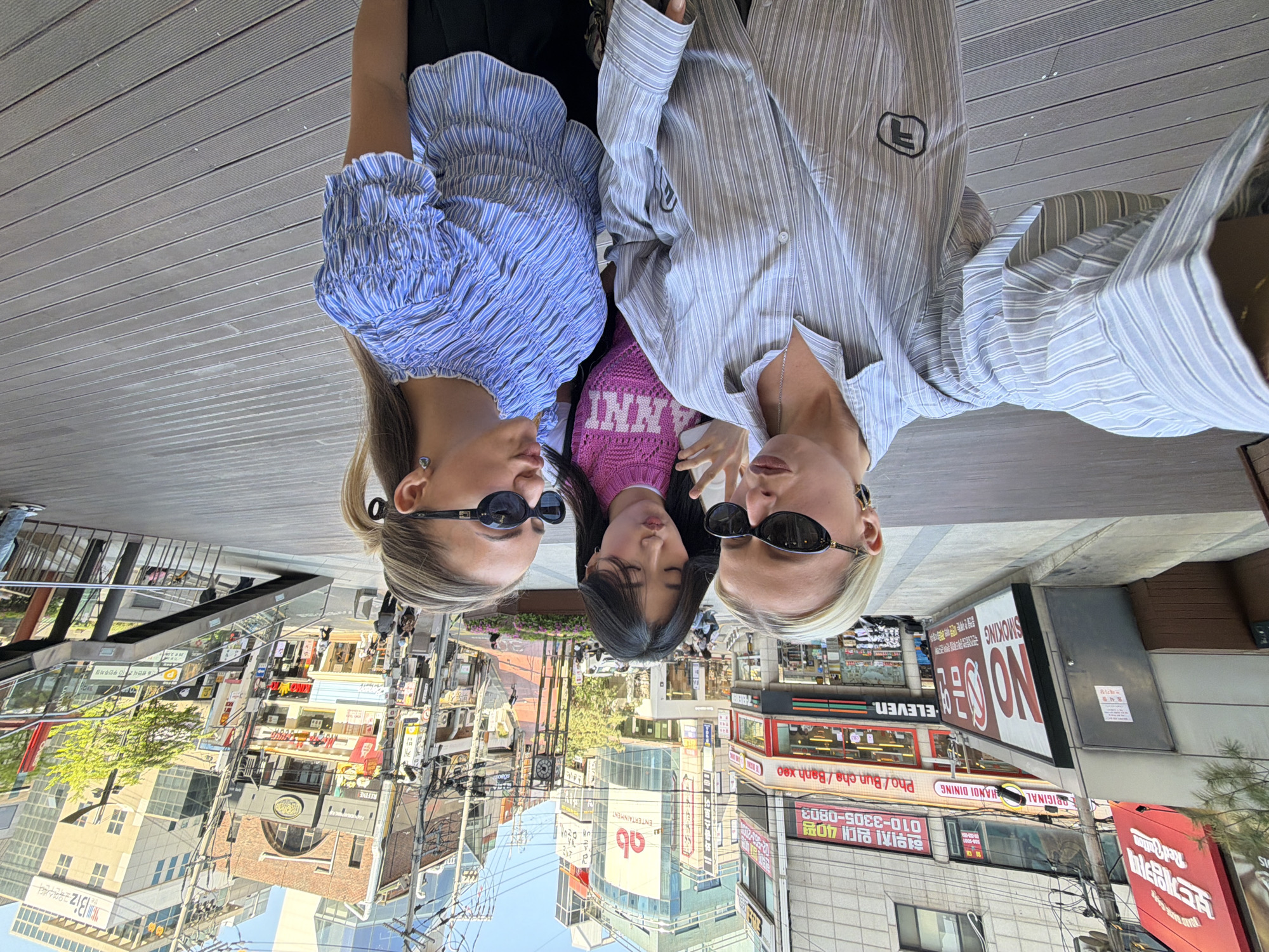 Here is a forensic description of the image:
**Scene Overview:**
The image appears to be a selfie taken by three individuals standing in a commercial area of Seoul, South Korea. The buildings and signs in the background give this indication. The viewpoint is downward, as if the photographer is holding the camera above the subjects.
**People:**
Three young women are centered in the shot.
* The woman on the left wears a blue striped blouse, dark sunglasses, and has her blonde hair pulled back into a ponytail.
* The woman in the center has dark hair and is wearing a pink sweater.
* The woman on the right wears a light gray and white striped shirt, dark sunglasses, and has blonde hair with dark roots.
**Objects:**
* Various commercial signs written in Korean and English are visible. These include signs for 7-Eleven, Vietnamese food, karaoke, entertainment, and an “ab” logo.
* Buildings include shops, restaurants, and what appears to be an entertainment venue.
* Power lines and telephone poles are visible among the buildings.
* The women are standing on a wooden boardwalk or platform.
* A small pink bag is carried by the central woman.
**Actions and Interactions:**
The women are posing together for the selfie, close together, with their faces near the center of the frame.
**Environment:**
The environment is a densely built urban area with a mix of commercial and entertainment venues. The sky is visible in the upper portion of the image and appears sunny. The ground is covered with what appears to be a boardwalk or platform. The presence of shops, signs, and infrastructure indicates a busy commercial area.
**Location (Seoul, South Korea):**
Based on the Korean language signage, building styles, and general urban setting, the location is highly probable to be in Seoul, South Korea.