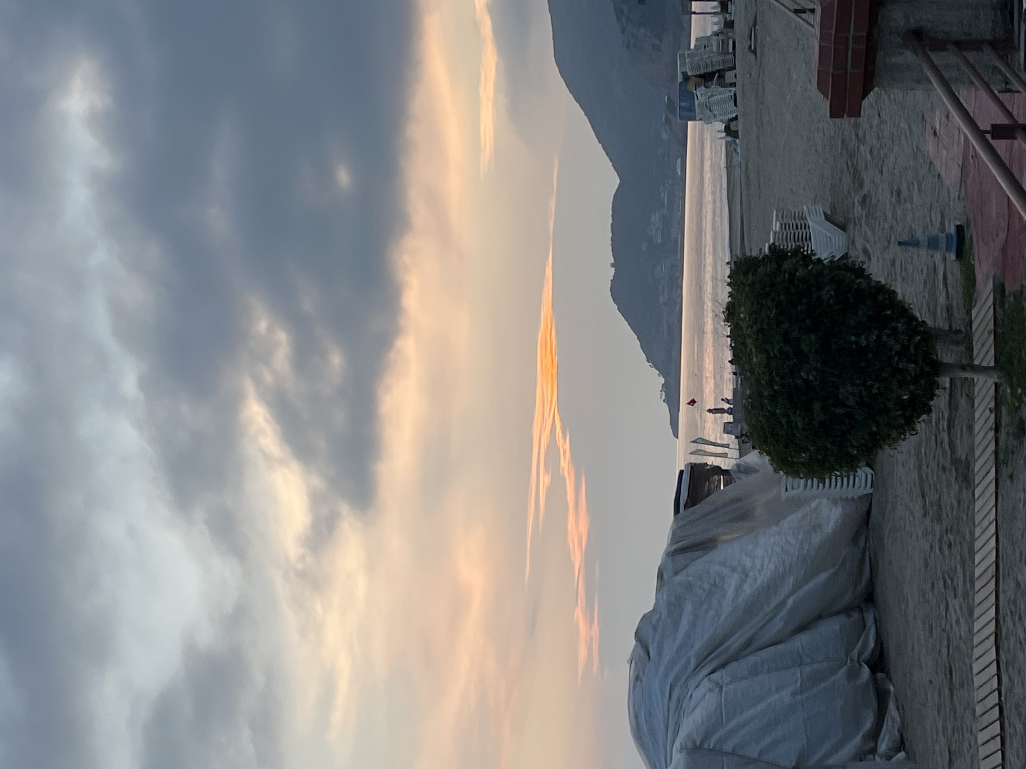 The image captures a serene coastal landscape at either sunrise or sunset, viewed in a landscape orientation (correcting for the original's 90-degree clockwise rotation). The sky, occupying the left and upper portions, is filled with varied clouds, ranging from dark grey to soft blues, illuminated by warm patches of orange and yellow light, indicating a low sun.
A dark, elongated mountain range stretches across the horizon, separating the glowing sky from the calm body of water below, which reflects the soft light with a silvery sheen. On the right, a sandy beach dominates the mid-ground and foreground. Numerous white or light-colored lounge chairs are stacked neatly on the sand, suggesting a resort or leisure area. A small, well-shaped, dark green bush or tree stands prominently on the beach. In the immediate foreground, a large, light-colored tarp is draped over an object, possibly covering furniture or equipment. A small blue cone is visible on the sand near the stacked chairs, and parts of a reddish-brown brick structure and a metal railing are seen on the far right.
Two small, indistinct human figures are visible near the water's edge in the distance, their specific activity not discernible. The overall scene is peaceful and tranquil. The low, warm light and the composition of mountains, sea, and beach are consistent with the coastal areas of Alanya, Turkey. There is no visible text in the image.