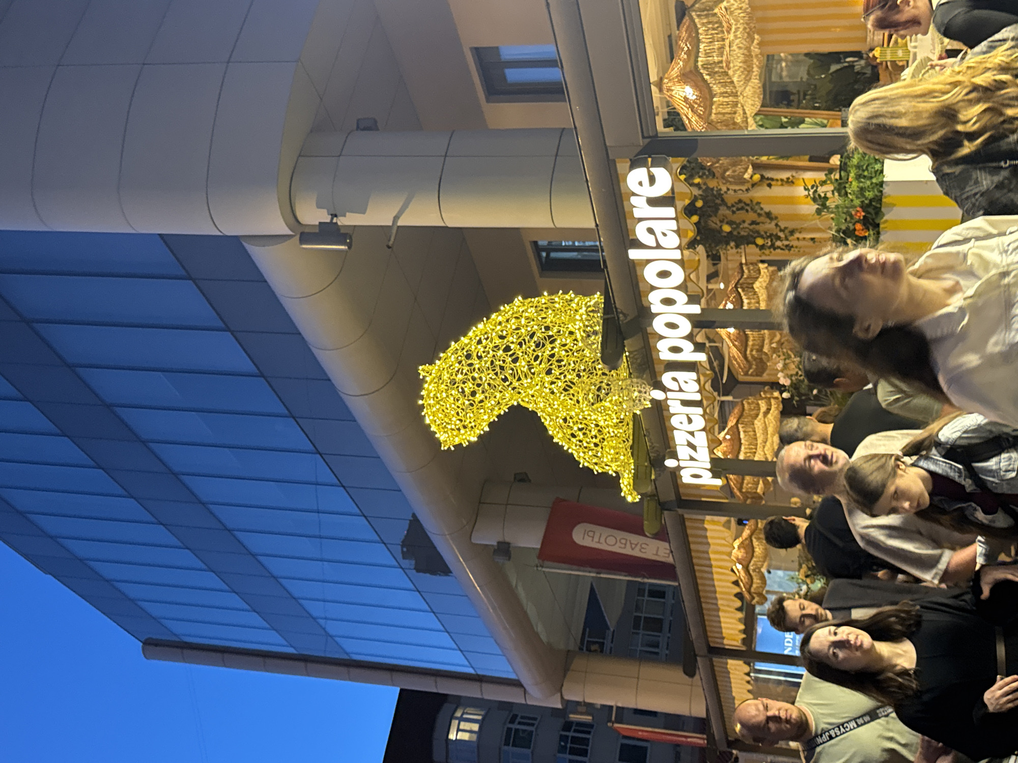A group of people are gathered outside a restaurant called "pizzeria popolare" in Yekaterinburg, Russia. The restaurant has a yellow and white striped awning and is decorated with yellow lights shaped like a bear. The building behind the restaurant is a modern office building with blue glass windows. The sky is dark blue, indicating it is evening.
