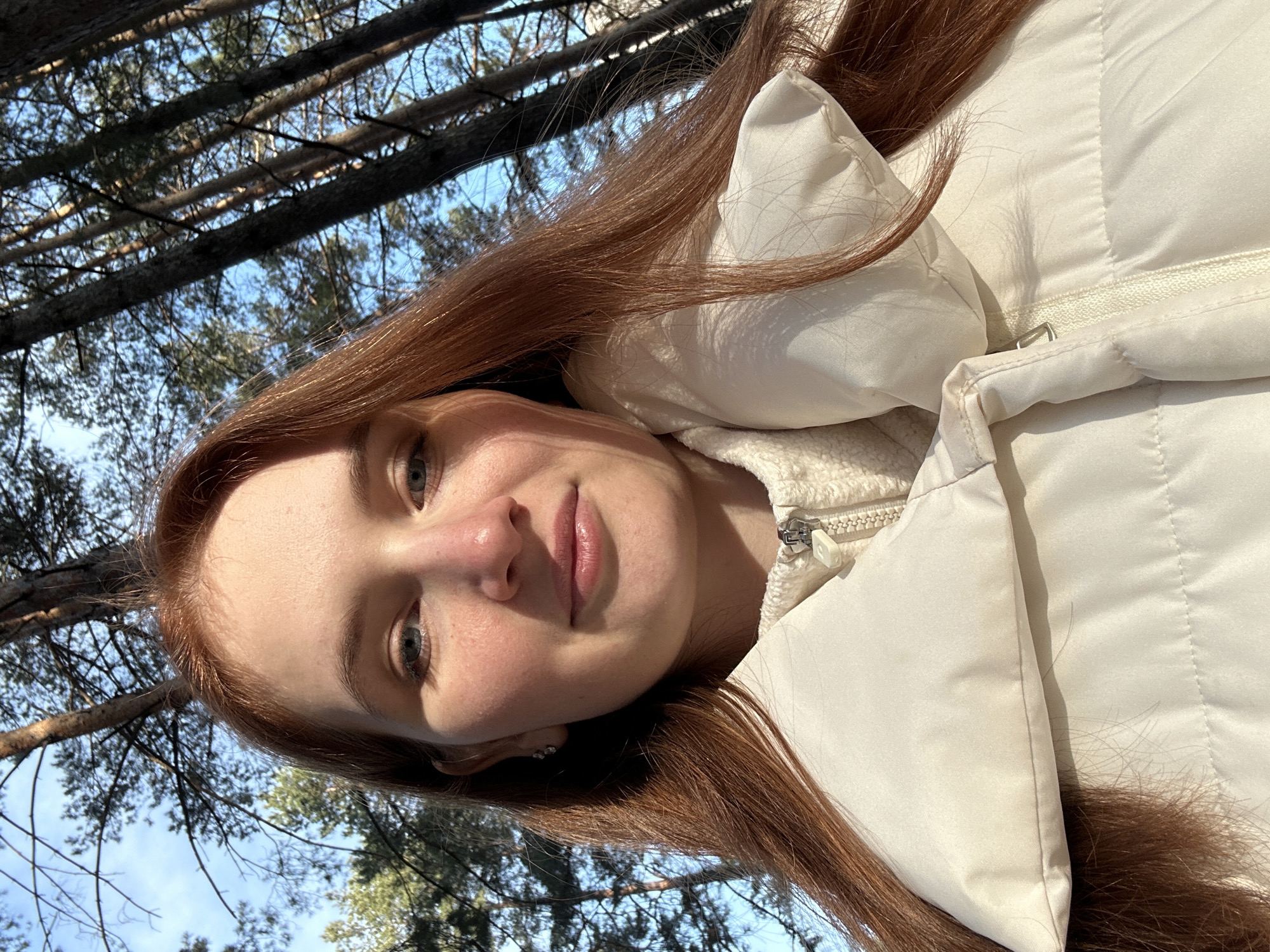 A young woman with long, reddish-brown hair takes a selfie in an outdoor setting. She wears a white puffy jacket. The background shows a forest with trees and a blue sky visible through the branches. The location is Chaikovsky, Russia.