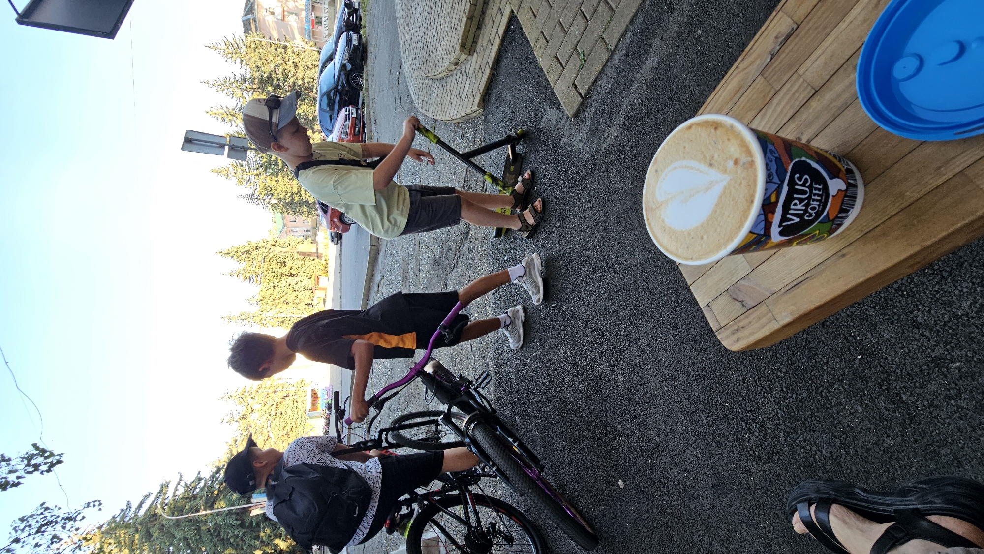 Three children are outdoors on an asphalt surface near a sidewalk with parked cars in the background. The child on the left, facing away from the camera, wears a black baseball cap and a backpack. They are next to a bicycle. The child in the middle, wearing a black and orange t-shirt and shorts, is holding the handlebars of the bicycle. The child on the right, wearing a light green t-shirt, shorts, and a baseball cap, is standing on a scooter. In the foreground, a cup of coffee with latte art is on a wooden table. A blue lid is to the right of the coffee cup. Trees and buildings are visible in the background.
