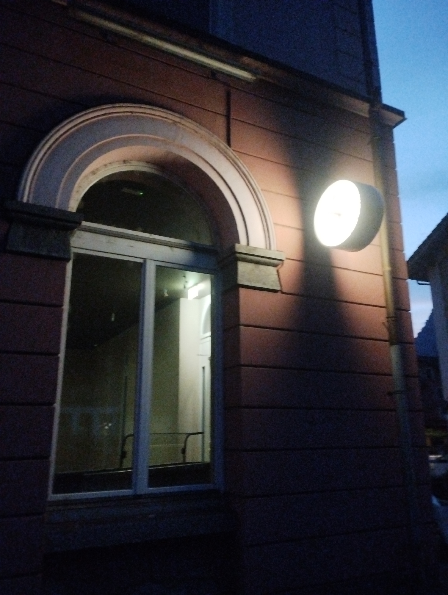 An exterior view of a building at dusk shows a large, arched window with a glass pane. The window is framed by a dark stone archway. A round, illuminated light fixture is mounted on the exterior wall to the right of the window, casting a shadow on the building's facade. The building's wall is constructed of dark, horizontal brickwork. The sky above is a deep blue, indicating twilight.