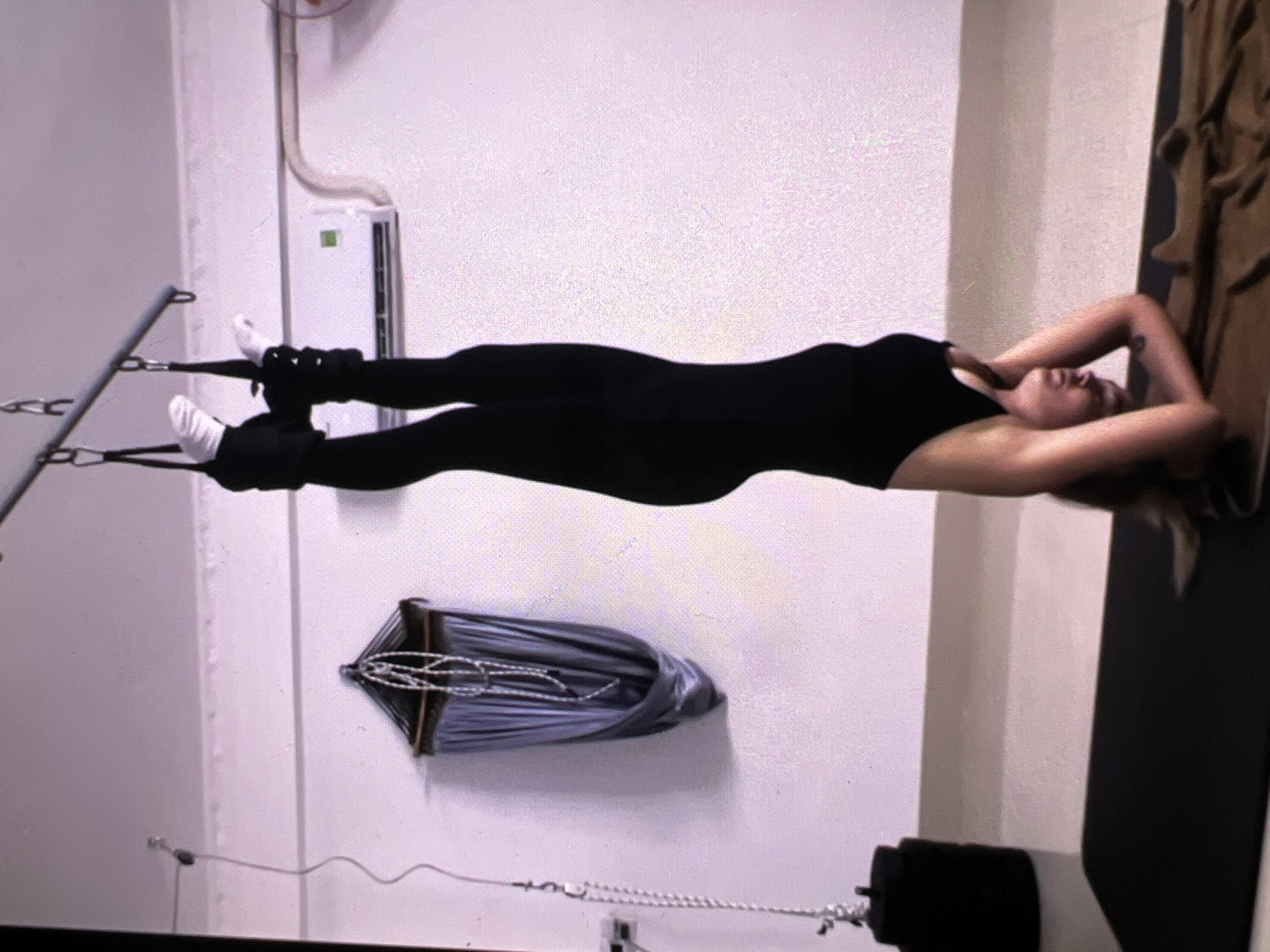 A person, likely a woman, is captured in a full-body shot, performing an exercise in an indoor fitness studio. She lies horizontally on her back, suspended or supported, with her head facing right and feet to the left. Her feet, wearing white socks, are secured in black ankle straps which are attached to a silver metal frame on the left side of the image. She is dressed in a form-fitting black outfit, possibly a leotard or activewear set. Her arms are bent, with hands clasped behind her head, and her face shows a relaxed expression with her eyes closed or looking upwards in concentration.
The setting is a room with plain white walls. Electrical conduits and wiring are visible on the upper left wall. Below the person's body, a folded grey or blue fabric, resembling a hammock or sling, hangs from the wall. A brown, textured object, possibly a blanket or towel, is partially visible on the far right. The activity appears to be a form of aerial fitness, Pilates, or core conditioning, utilizing specialized suspension equipment. No visible text is present in the image.