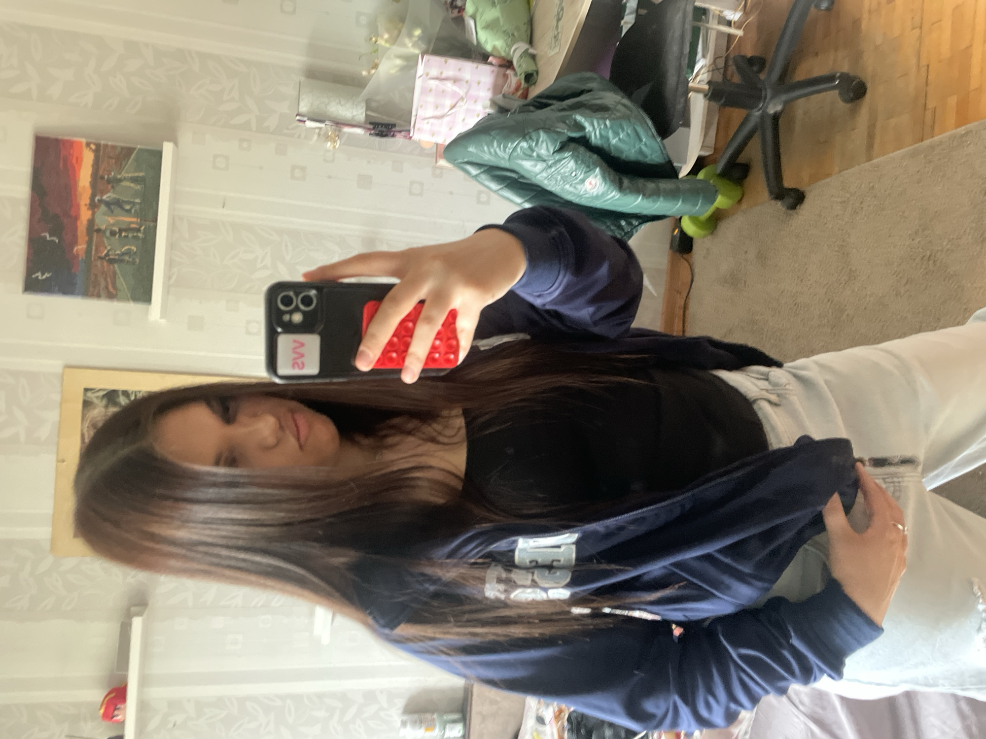 A mirror selfie captures a young woman in an indoor setting. She holds a black smartphone with a red pop-it grip on the back, visible in her right hand. On the phone case, reversed text "2VV" is seen on a white sticker. The woman has long, dark brown hair and a neutral expression, looking towards the camera. She is wearing a navy blue zip-up jacket, under which a black top is visible, and light-colored jeans. White letters, possibly "EE" or "LE", are partially discernible on the left chest of her jacket.
The background reveals a room with light-colored wallpaper featuring a subtle leaf pattern. To the left, a painting hangs on the wall, depicting dark figures against a vibrant orange and red sunset sky, with two small white birds. Various objects are scattered around, including a green puffy jacket draped over a surface, and the base of a black office chair with wheels. The floor is carpeted in a light beige color. The overall setting suggests a casual home environment in Vsevolozhsk, Russia. The time of day is not evident.