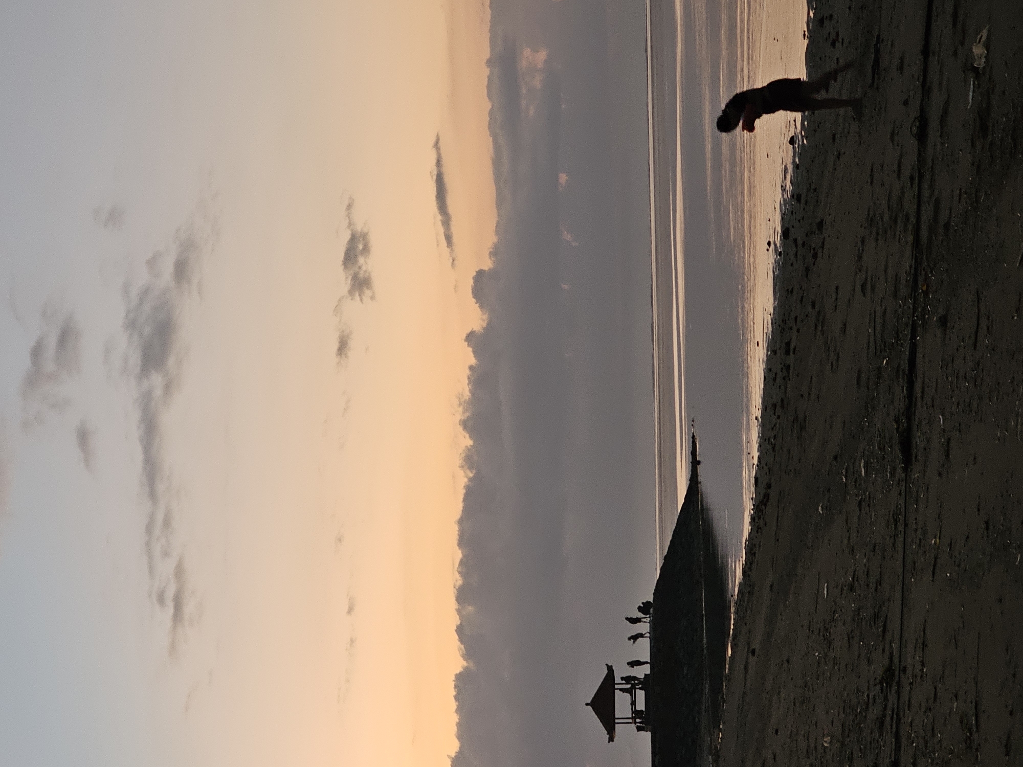 This image depicts a tranquil beach scene at sunrise or sunset in Denpasar, Indonesia. The sky transitions from a soft orange near the horizon to a pale blue overhead, with scattered clouds in varying shades of gray and white. In the mid-ground, silhouetted figures are visible near a traditional Balinese gazebo situated on a small, dark hill. Several individuals are standing around and inside the gazebo, while others are walking or sitting nearby. The calm ocean water reflects the colors of the sky. In the foreground, a sandy beach is partially visible, with the dark silhouette of a single person in motion on the right side of the frame. The overall atmosphere is serene and peaceful.