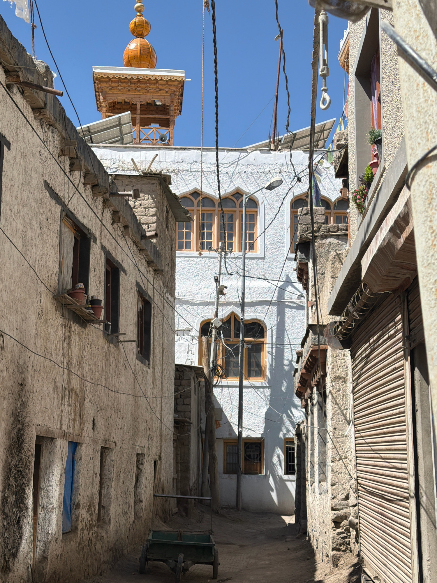 The image depicts a narrow alleyway in the old city of Leh, India, with traditional Ladakhi architecture. The alley is flanked by two- and three-story buildings constructed from mud brick and stone, with rough-textured walls and wooden window frames. The building on the left has several small windows and a wooden ledge with potted plants. The building on the right features a large, corrugated metal roller door.
In the background, a white building with arched windows and wooden frames is visible. Atop this building, a wooden structure with a golden dome and intricate carvings stands against a clear blue sky. Solar panels are installed on the roofs of some buildings. Electrical wires and poles are a prominent feature, crisscrossing the scene.
In the foreground, a small, green, two-wheeled cart with a wooden handle rests on the dusty ground. The alleyway is unpaved and shows signs of wear and tear. The overall impression is one of a well-preserved, historic urban environment bathed in bright sunlight.