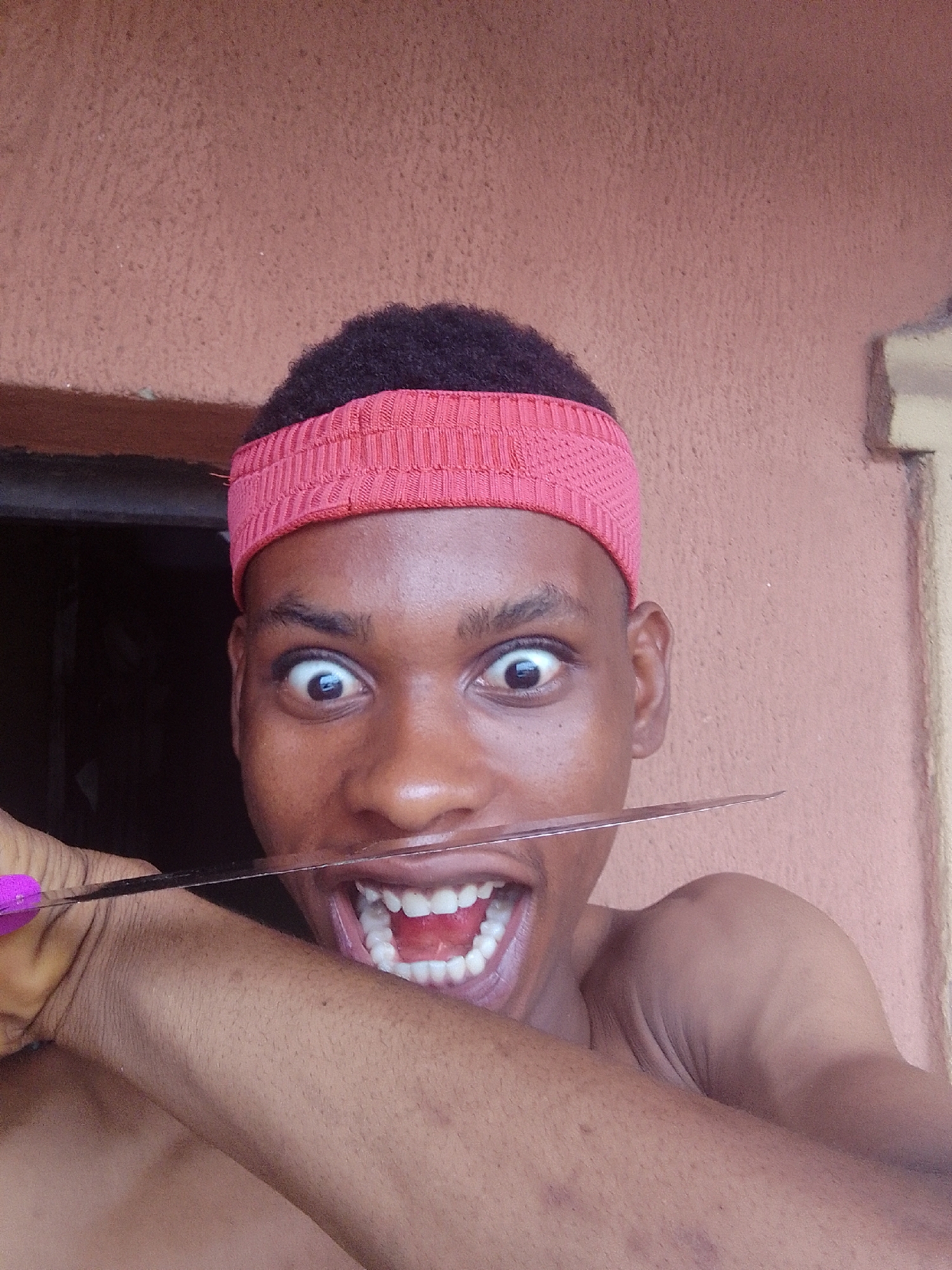 A close-up portrait captures a young person with dark skin, short dark hair, and a red knitted headband. The person is positioned centrally, looking directly at the camera with wide, exaggerated eyes. Their mouth is open in a very wide smile or grimace, revealing a full set of bright white teeth and their tongue.
A long, thin, metallic blade is held horizontally across their mouth, just below their nose and above their lower lip. The blade extends from one side of their face to the other. The handle of the blade, partially visible on the left, is purple and is held by the person's left hand, with a thumb visible. The person's bare shoulder and arm are visible in the foreground, below their head.
The background consists primarily of a textured, reddish-brown wall. To the left of the person's head, a dark, rectangular opening, possibly a window or doorway, is visible. A lighter, cream-colored section of a wall is present on the far right. The overall expression of the person is highly theatrical, conveying a sense of exaggerated surprise, intensity, or playfulness. No text is visible in the image.