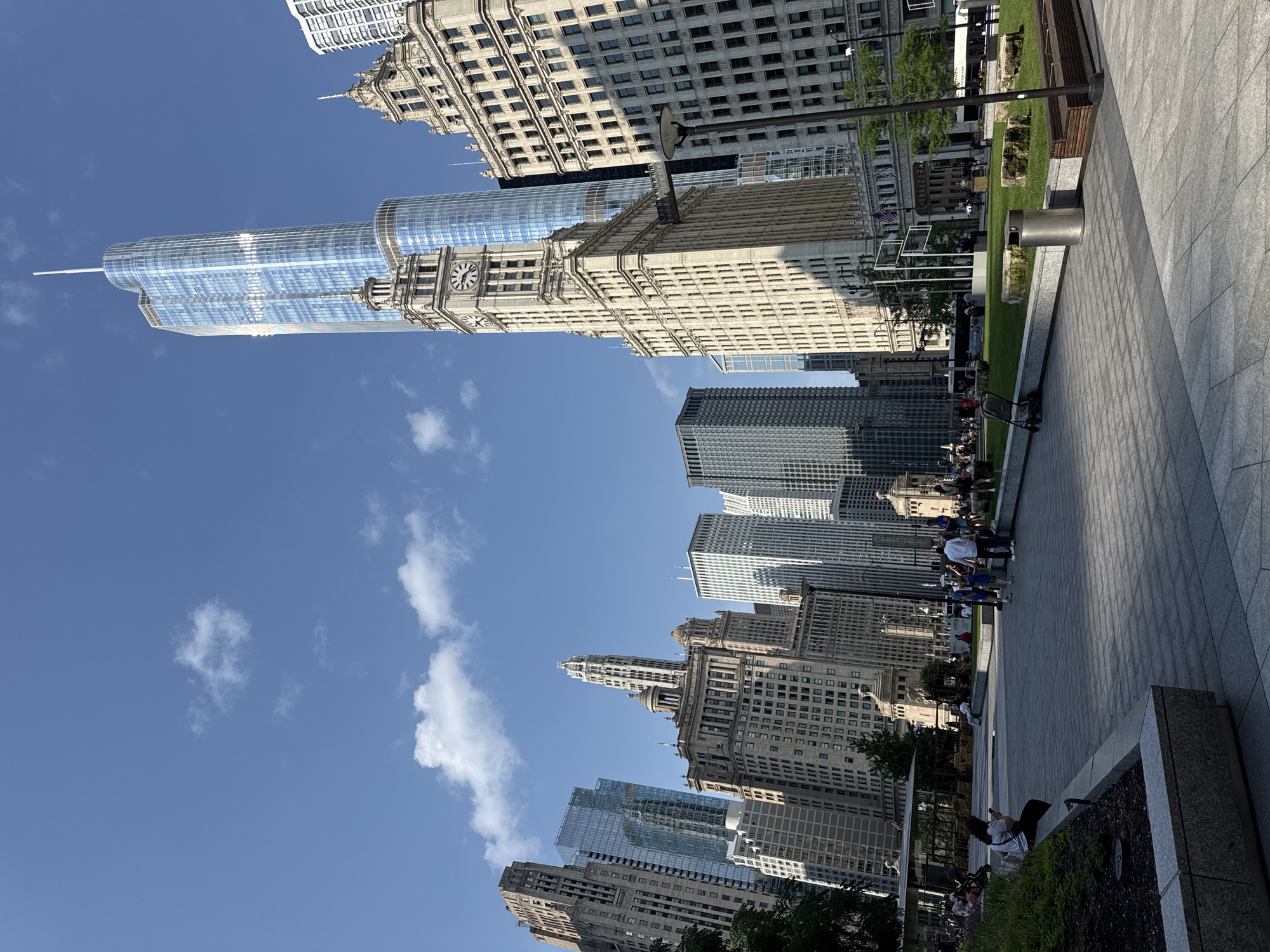 The image depicts a sunny day in Chicago, Illinois, USA, showcasing a cityscape with a prominent park area in the foreground. Tall, modern skyscrapers with glass facades and older, ornate buildings with stone facades dominate the background. A distinctive clock tower stands out on one of the older buildings. The park features manicured green lawns, stone benches, paving stones, and some decorative elements. Several people are visible in the park, some walking, others sitting or standing, engaged in various leisurely activities. The sky is blue with scattered white clouds. A bus and a trash can are also visible within the park.