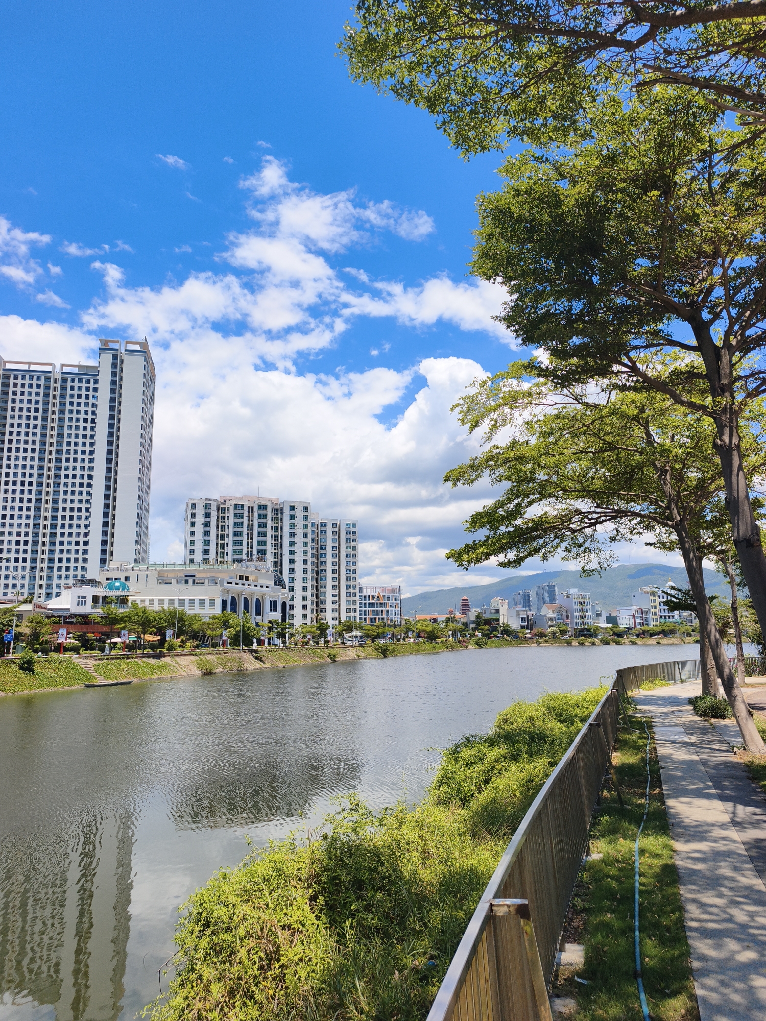 The image depicts a waterfront scene in Quy Nhon, Vietnam, likely along the Thi Nai Bay area or a river within the city. On the left bank, a row of modern, tall white apartment buildings with numerous windows dominates the skyline. In front of these buildings, a lower, white structure with arched windows and a covered walkway is visible. A street lined with trees and some smaller buildings runs along the waterfront.
The river in the foreground is calm, with reflections of the buildings and the sky visible on its surface. A paved walkway runs along the right bank of the river, bordered by a metal fence. Lush green vegetation and trees line the edge of the walkway, casting shadows on the path. The sky is a bright blue with scattered white cumulus clouds, suggesting fair weather. In the distance, more buildings and rolling hills can be seen. There are no people visible in the image.
