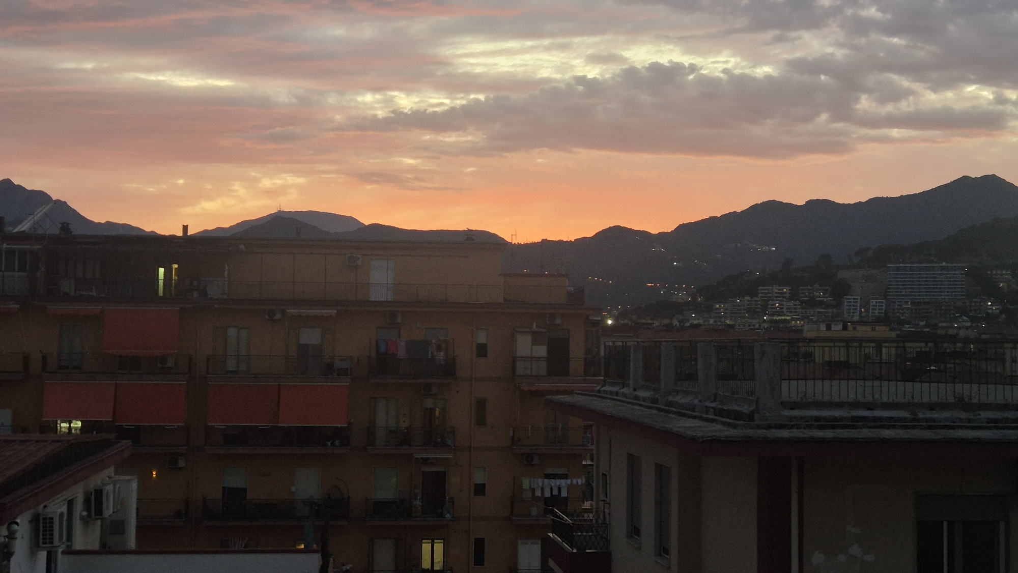An outdoor medium shot captures a city skyline at dusk, with mountains forming the backdrop and a vibrant orange and pink sky overhead. In the foreground, a multi-story building with an orange facade is visible, featuring balconies with awnings and laundry hanging out to dry. Another building with a red roof is partially seen on the left. In the middle ground, more buildings with lights on are scattered across the landscape, and in the background, a large mountain range is silhouetted against the colorful sky. The scene appears to be an urban residential area at the foot of a mountainous region.
