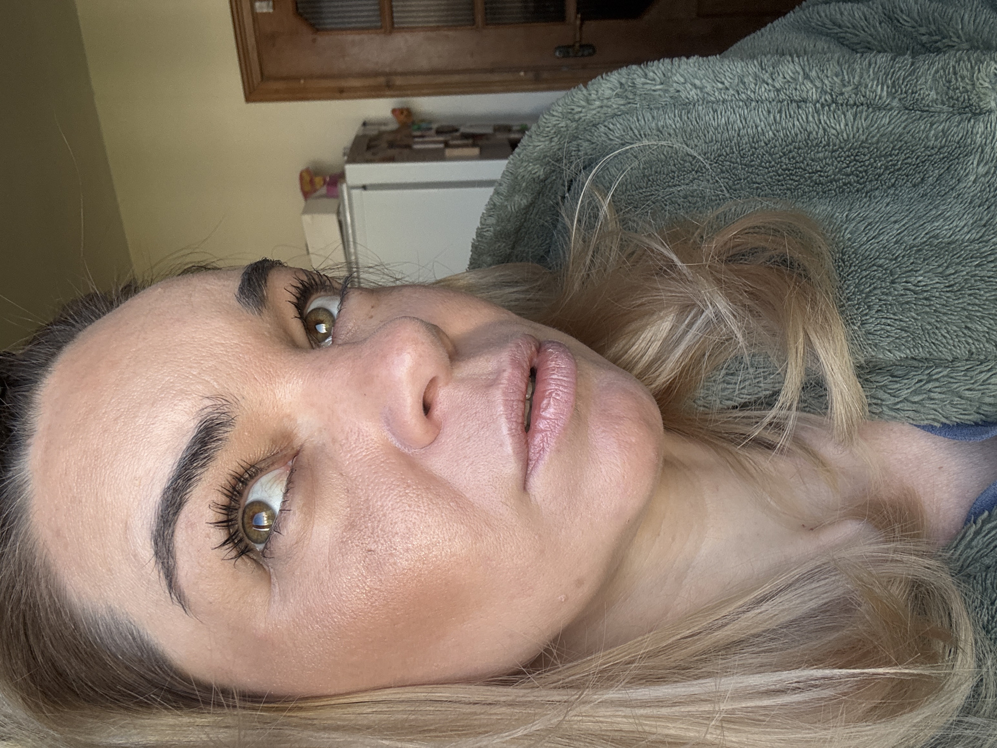 The image is a close-up, slightly angled portrait of a woman with light skin and blonde hair. She is wearing makeup, including mascara and lip color. Her eyes are open and looking upwards. A portion of her body is covered by a green, fuzzy jacket or blanket. The background includes a white cabinet and a wooden cabinet above it.
There is no indication of the location being in the city of Novoe Devyatkino, Russia.