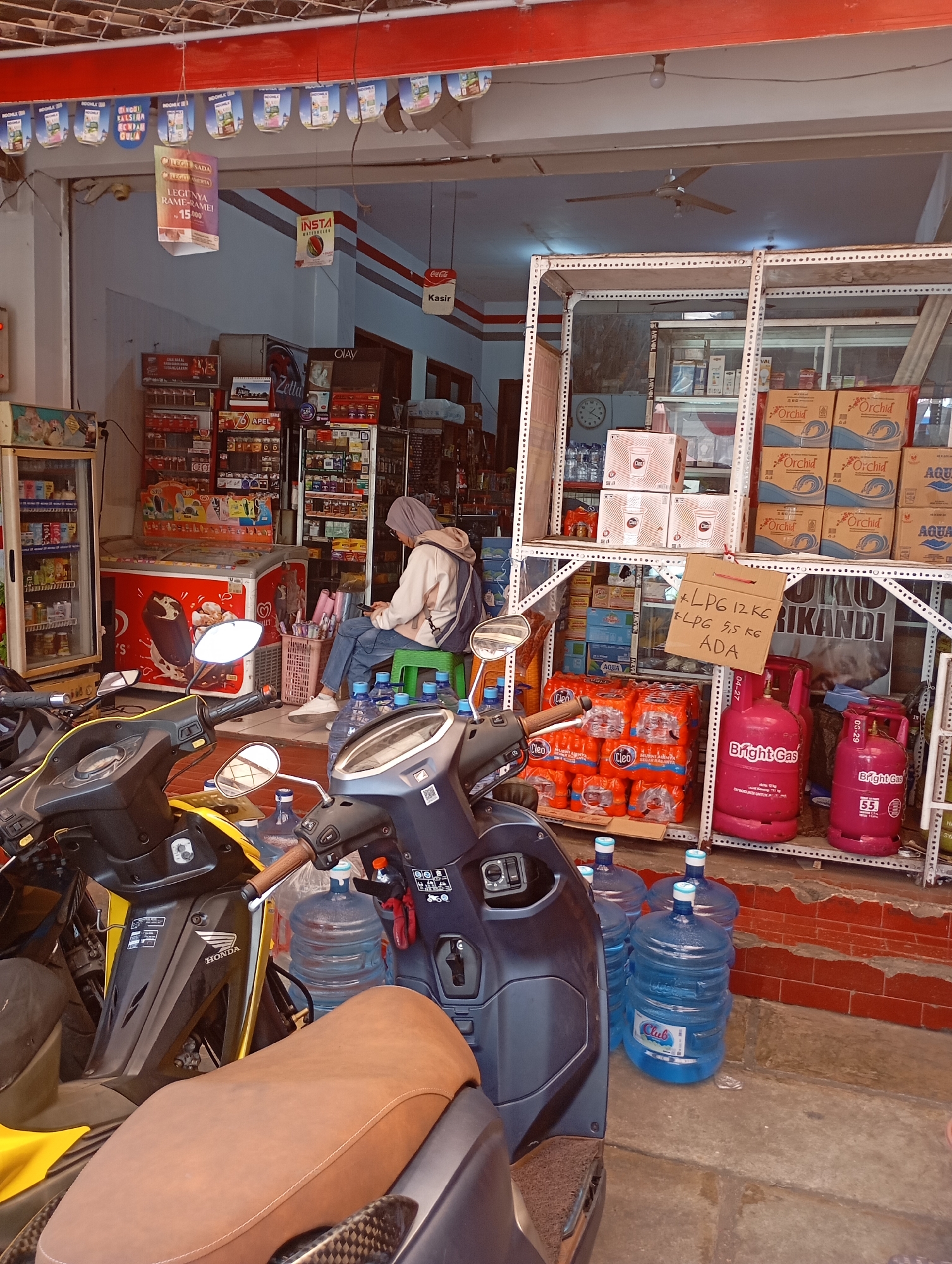 The image depicts an outdoor convenience store or small shop in Malang, Indonesia, likely a "warung" or "toko kelontong." The scene is cluttered and bustling with goods. In the foreground, several motorcycles are parked, indicating this is a common mode of transport for customers. The shop itself appears to be open to the street, with shelves stocked with various items including bottled water, cooking gas cylinders, snacks, and possibly household products. A young woman wearing a hijab and a light-colored hoodie is seated on a green stool, looking at her phone. She is positioned near the entrance, amidst stacks of goods. Above the entrance, decorative banners and signage are visible, some advertising products. A red Coca-Cola sign is also present. The overall impression is of a busy, typical retail establishment in a Southeast Asian city, catering to local needs.