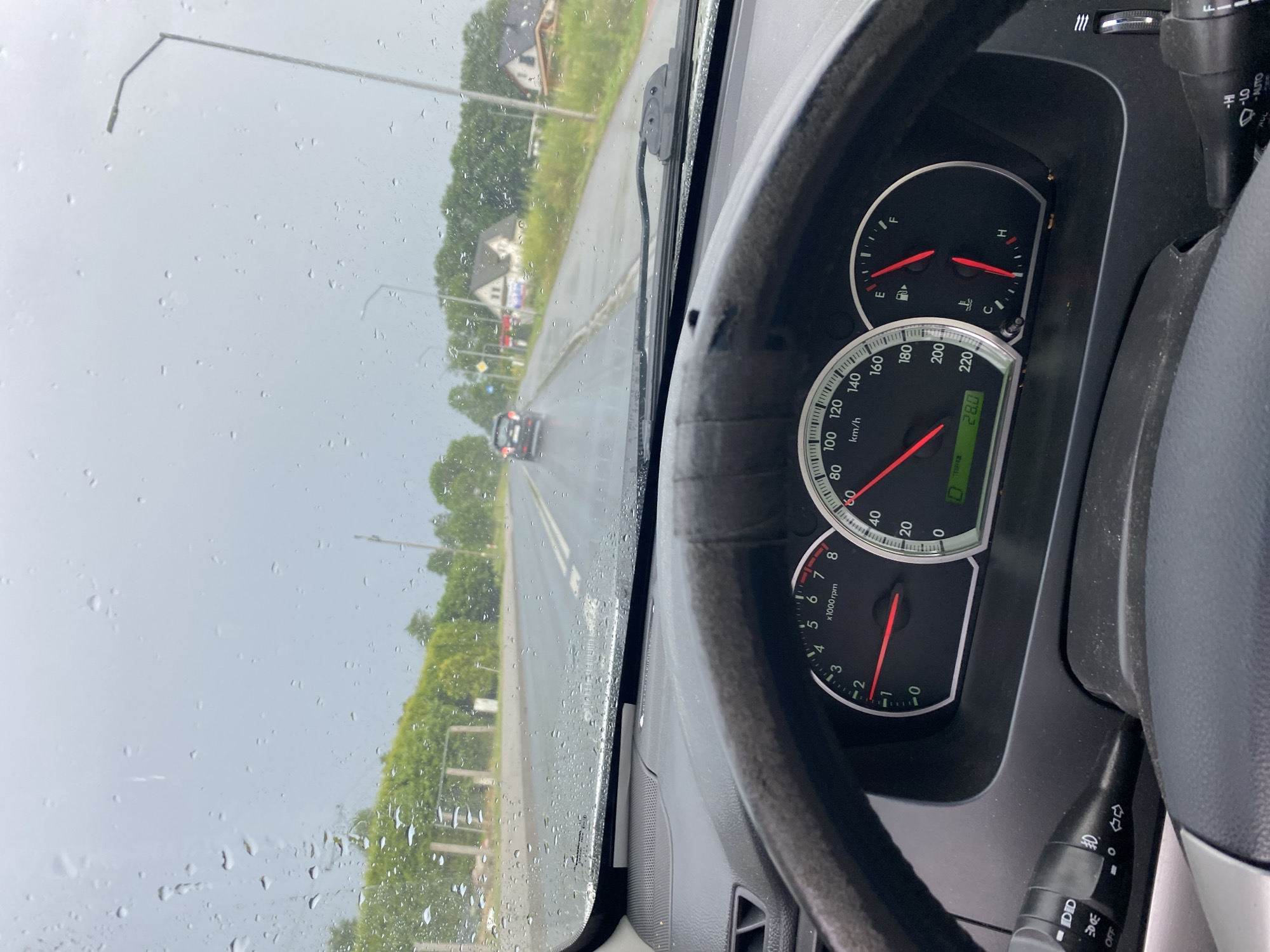 The image is taken from inside a car, looking out through a rain-streaked windshield. The perspective is from the driver's seat, with the dashboard and steering wheel visible on the right side of the frame. The dashboard displays a speedometer that reads approximately 28 km/h, and a fuel gauge. Outside the car, it appears to be daytime with overcast skies, suggesting rainy or stormy weather. The road is wet, and trees line the side of the road. In the distance, there are houses and a street light. The scene is set in Przęsocin, Poland. The overall impression is of driving in inclement weather.