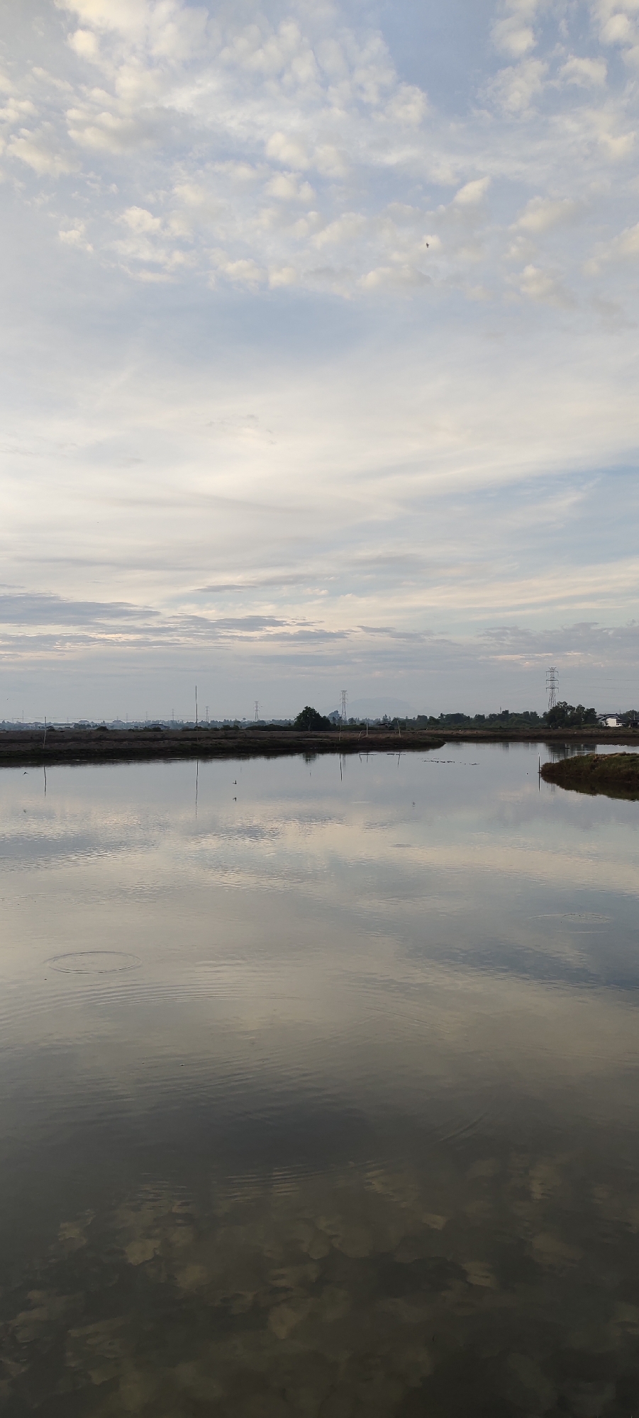 This is a landscape photograph taken in Lhokseumawe, Indonesia, depicting a calm body of water with reflections of the sky and surrounding landscape. The sky is filled with a mix of scattered clouds and wispy cirrus clouds. In the distance, a treeline is visible, punctuated by several tall electrical pylons. The water's surface is smooth, creating clear reflections of the sky and the faint outlines of structures and vegetation along the shore. The lower portion of the image shows the shallow, murky bottom of the water, with some indistinct shapes visible beneath the surface. There are no people or animals clearly visible in the image. The overall impression is one of a tranquil, rural or semi-rural setting at what appears to be dawn or dusk.