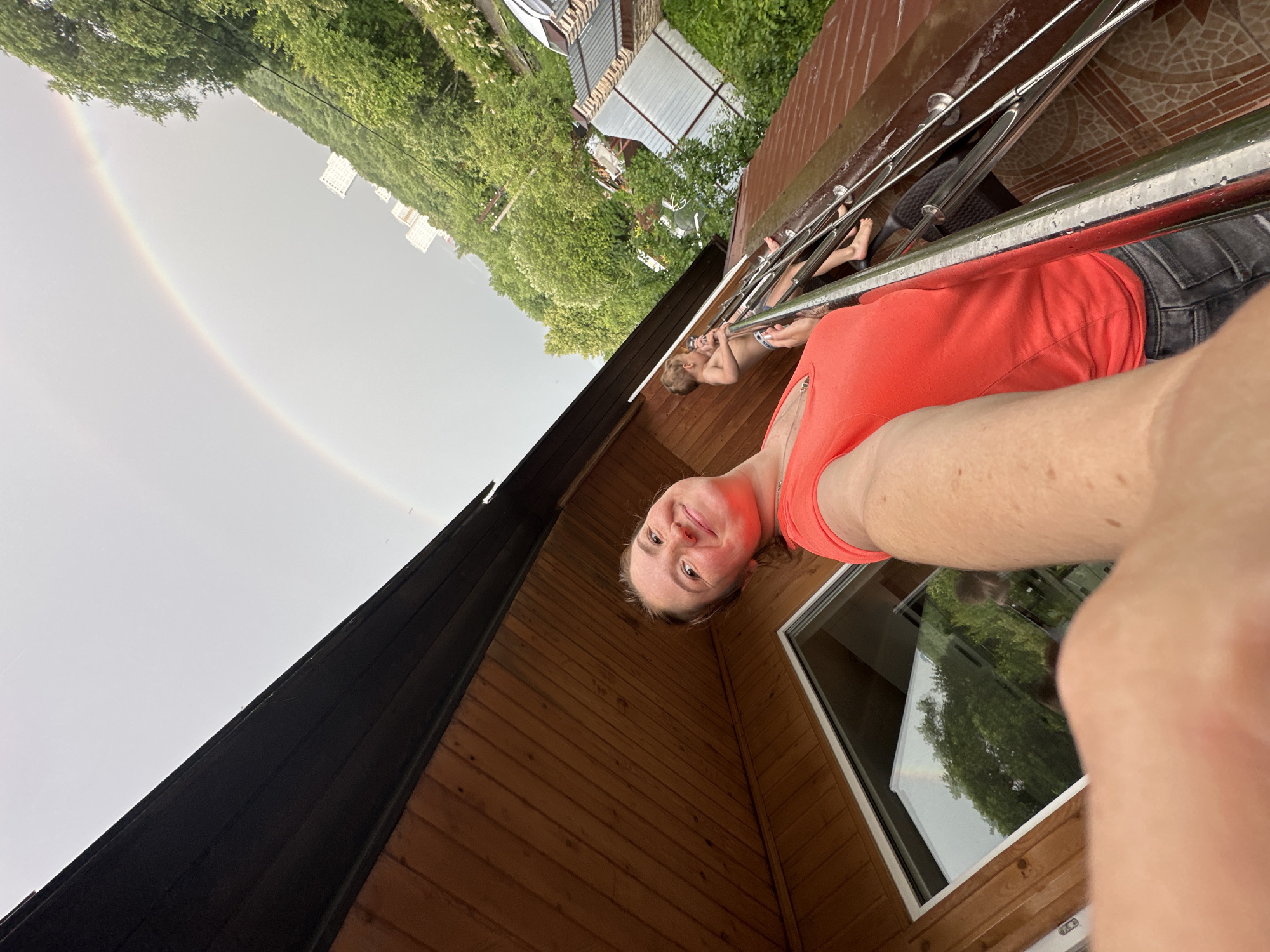 A selfie taken from a low angle shows a woman with a smile, looking towards the camera. She is wearing an orange tank top and dark jeans. Her arm is prominently in the foreground. Behind her, a wooden structure with a dark roof is visible, as well as a window with a reflection of trees and a rainbow in the sky.
In the background, several children are visible on a metal railing or structure. One child, a young boy, is sitting and holding onto the railing, wearing swim trunks. Another child's legs are also visible. The setting appears to be outdoors, possibly a backyard or a porch area. Green trees and some buildings are visible in the distance. A faint rainbow arches across the sky, suggesting it may have recently rained. The scene suggests a casual, possibly summer, day. The location is identified as Nizhny Novgorod, Russia. There is no visible text in the image.