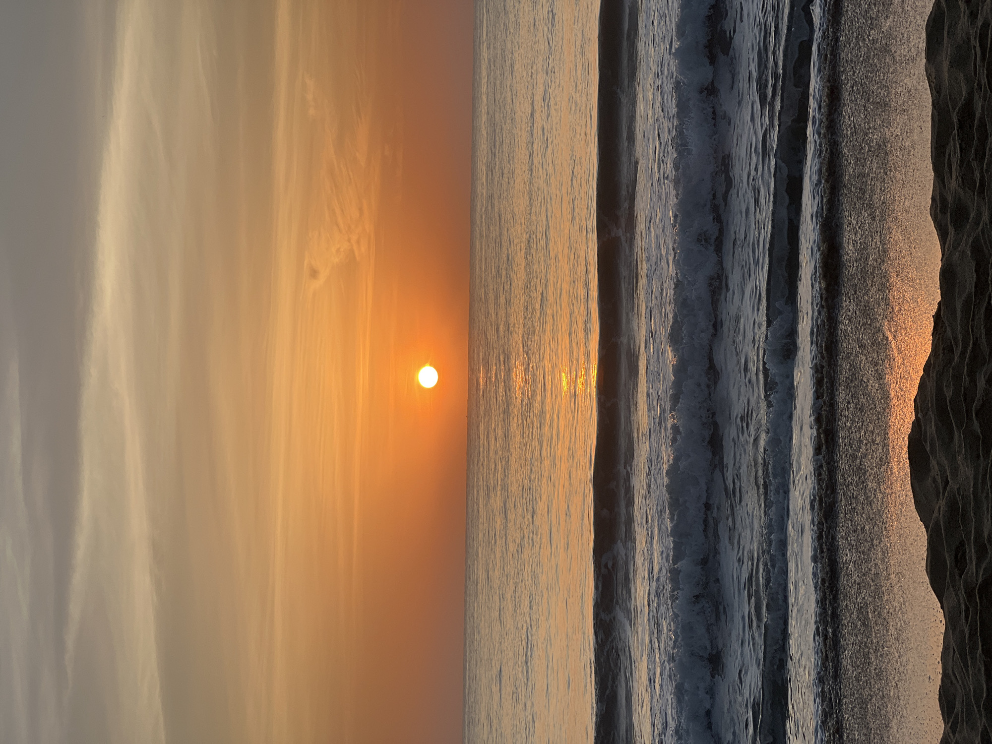 This is a vertical, eye-level, full shot of a beach at sunrise in Panama, Sri Lanka. The sun, a bright orange orb, is positioned in the center of the frame just above the horizon line, casting a shimmering golden reflection on the calm ocean water. Several layers of small to medium waves are breaking closer to the shore, creating white foam that washes onto the wet sand. The sky transitions from a pale gray at the top to a vibrant orange near the horizon, with thin wispy clouds scattered across the upper portion. The foreground consists of dark, textured sand, with dunes visible at the very bottom of the frame. There are no people or animals present in the image.