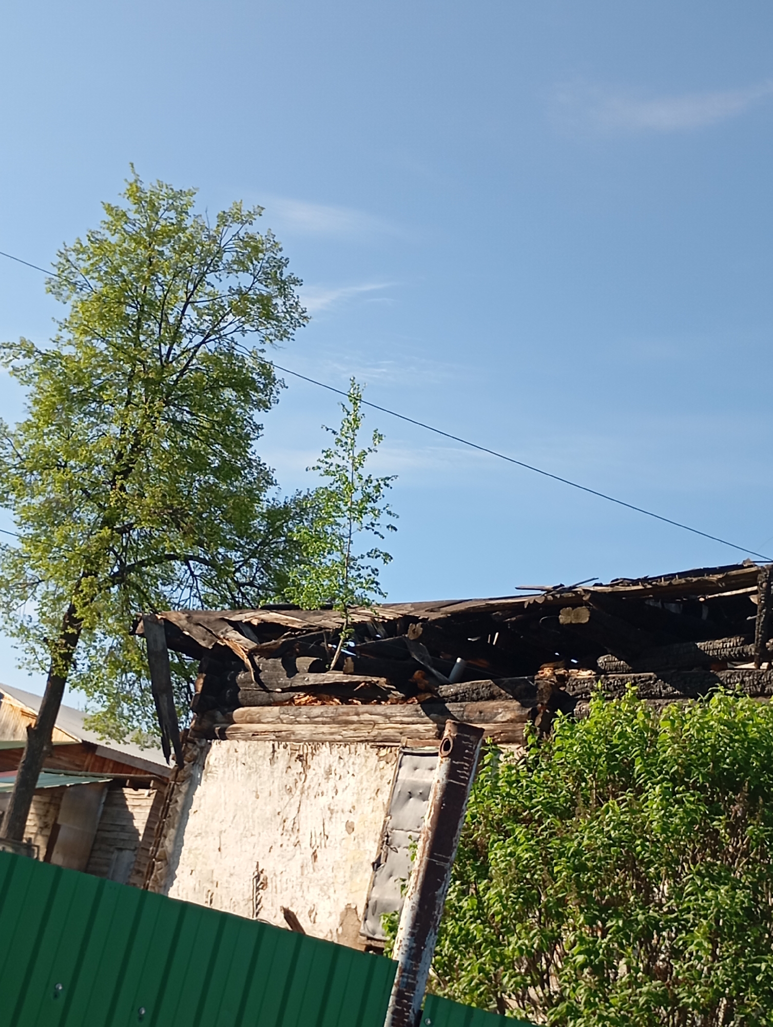 The image depicts a partially collapsed building in an urban area. The building appears to have been damaged, possibly by fire, as evidenced by the charred wood and missing sections of the roof. A portion of the building's facade is visible, with a dilapidated white exterior and exposed wooden beams. Adjacent to the damaged structure, there is a green fence and vegetation, suggesting a residential or suburban setting. Overhead, a clear blue sky and a power line can be seen. A tree stands near the building. The location is in the city of Unknown, Unknown.