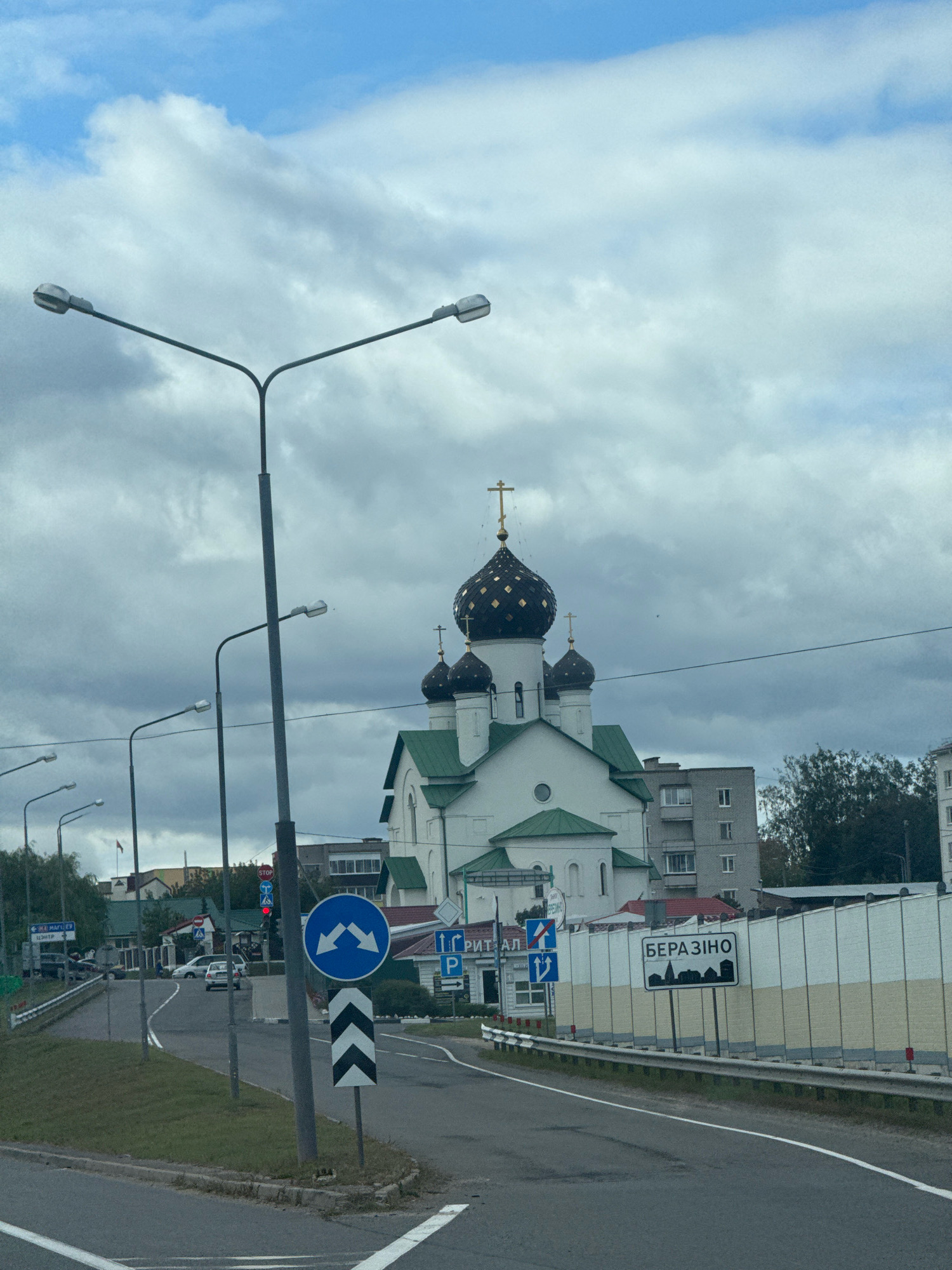 The image shows a street scene in Byerazino, Belarus, featuring a prominent white church with dark domes and a gold cross on top. The church has green accents on its roof. Several streetlights line the road, which curves through the scene. Various traffic signs are visible, including a "STOP" sign, directional arrows, and a sign with the city name "БЕРАЗІНО" (Byerazino) and a silhouette of buildings. Buildings of different sizes and architectural styles are present, including apartments and smaller structures. The sky is overcast with some blue patches and clouds. The road has white markings and a grass median. A white protective barrier runs along the side of the road. There are no people or vehicles clearly identifiable in the foreground.
