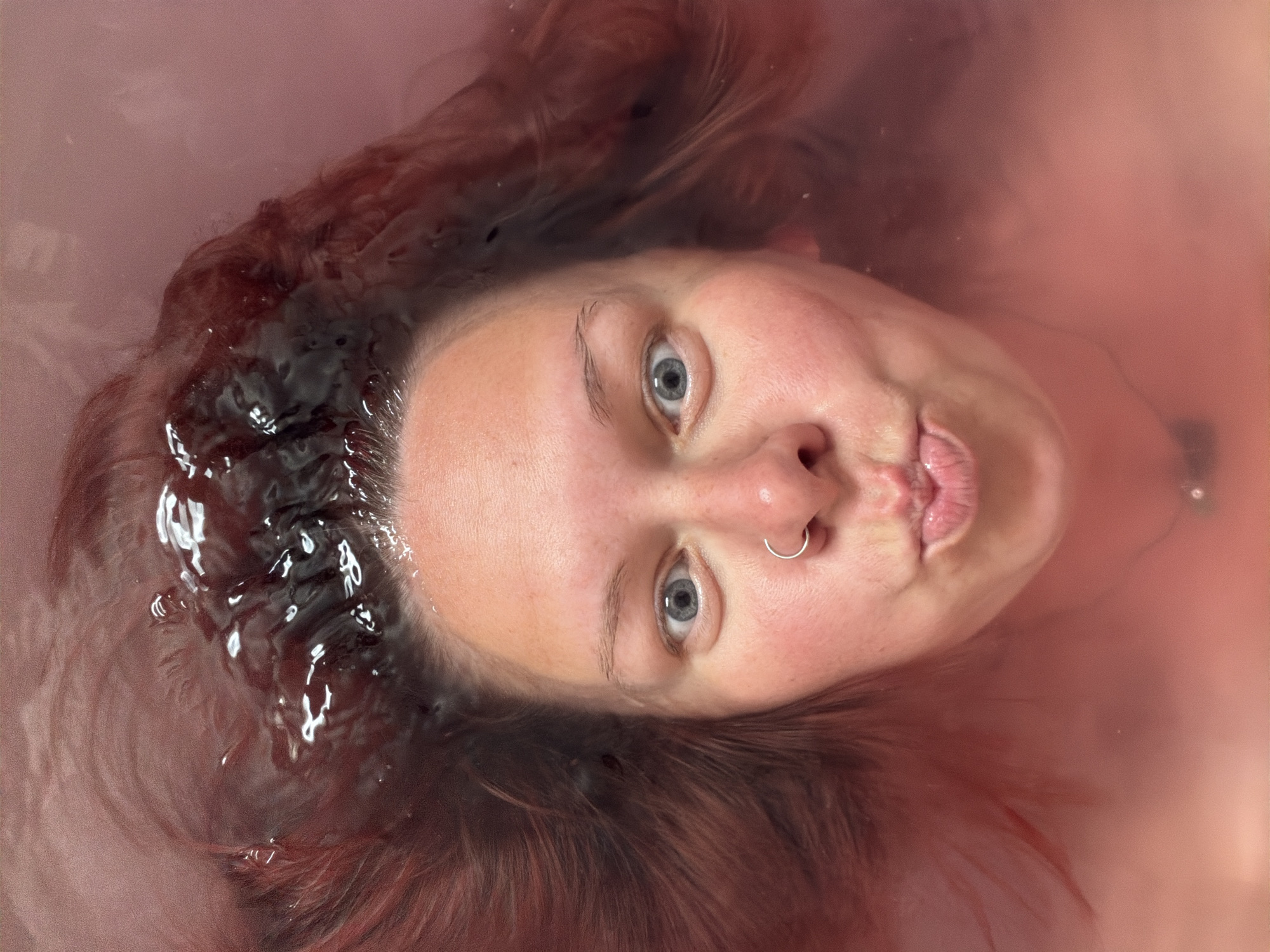 A woman floats on her back in water, her head the focal point. Her hair is partially submerged, creating a dark halo around her forehead. She has fair skin, light blue eyes, and a silver nose ring. Her lips are pursed. The water is a reddish-pink hue. The location is indeterminable from the image. The city of Chaikovsky, Russia cannot be verified from this image.