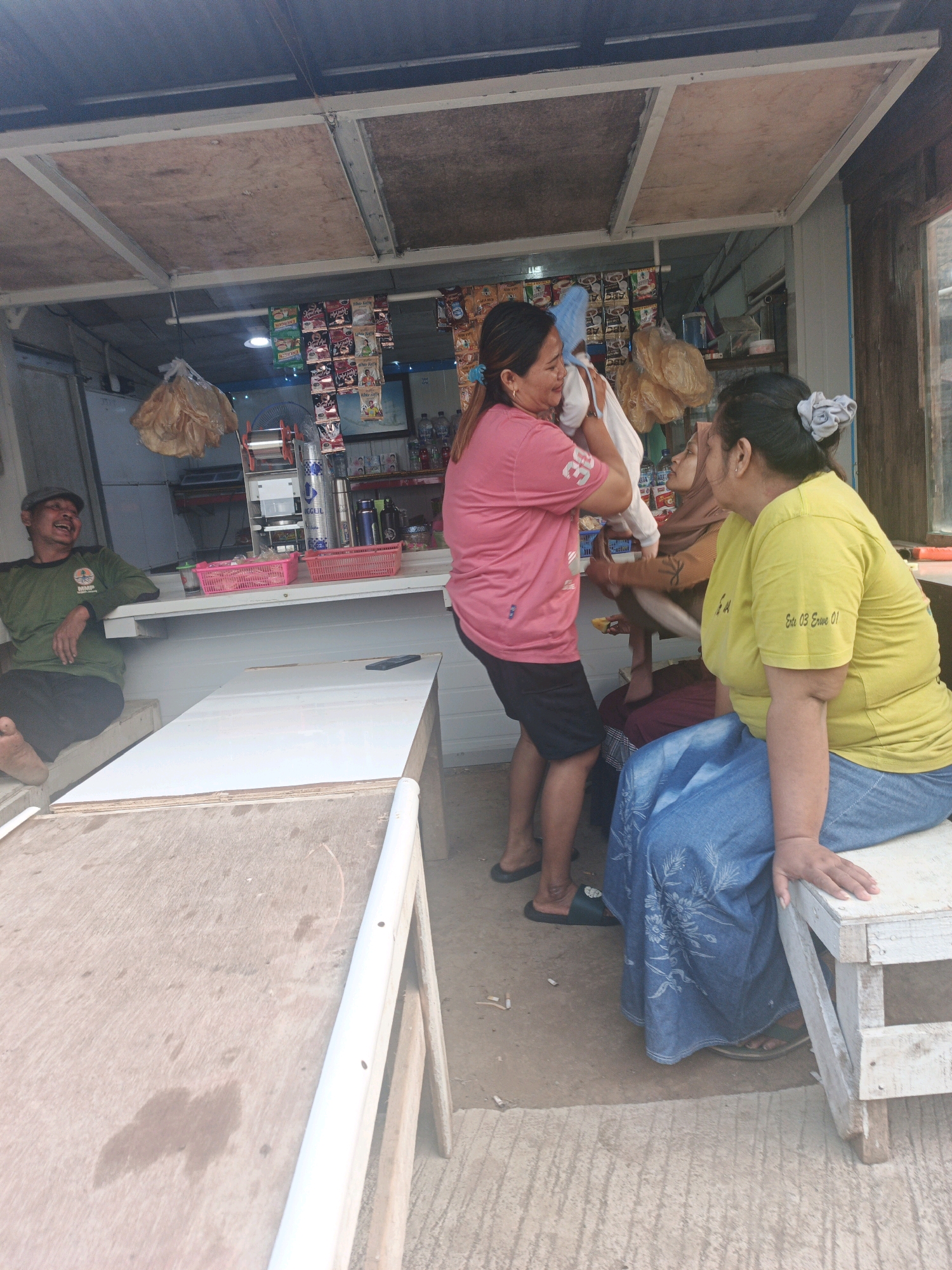 The image depicts an outdoor scene at a small, open-air stall in Unknown, Indonesia. In the foreground, two wooden tables are visible, the one on the left with a rough wooden surface and the one in front with a white laminate top. A man wearing a green t-shirt and a grey cap is seated on a bench to the left, laughing. Behind him, a counter is laden with goods, including pink plastic baskets filled with items, bottles of water, and packets of snacks displayed on shelves. Two women are engaged in an interaction at the counter. One woman, wearing a pink t-shirt and dark shorts, is holding a white object, possibly merchandise, and looking towards another woman. The second woman, wearing a yellow t-shirt and a blue patterned skirt, is seated on a white bench and looking at the first woman. A third woman, wearing a brown hijab and a dark top, is partially visible behind the counter, between the two women. The stall itself is constructed with a wooden roof and metal supports. The ground is a mix of concrete and dirt. Sunlight filters through the roof, creating a bright atmosphere.