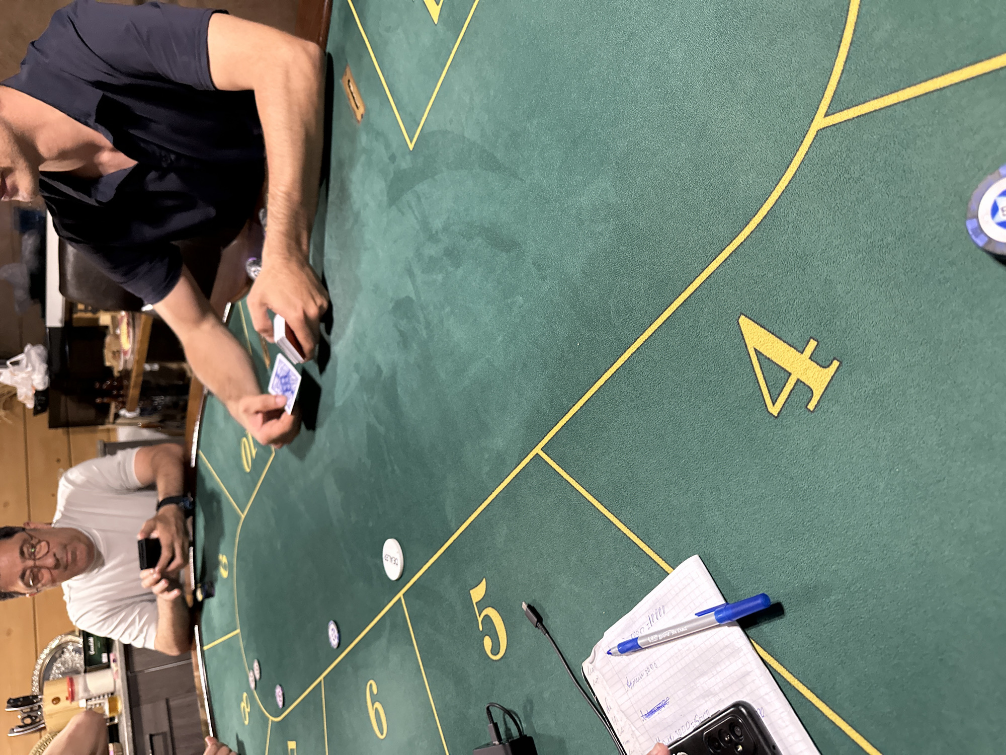 The image depicts a top-down view of a green felt poker table in what appears to be a residential setting. The table is marked with yellow lines and numbers, indicating seating positions or game progression. Several people are visible playing poker. In the foreground, a hand holds a blue pen and a cell phone, resting on a piece of paper with handwritten notes. A black charging cable for the phone is also visible. Further into the scene, a man in a white t-shirt and glasses is holding a phone. To his right, another man in a dark polo shirt is holding a deck of cards. There are poker chips scattered on the table. The background shows a counter with various items and wooden walls, suggesting a home environment. The lighting is dim, creating a casual atmosphere. The location is specified as Krasnaya Polyana, Russia.