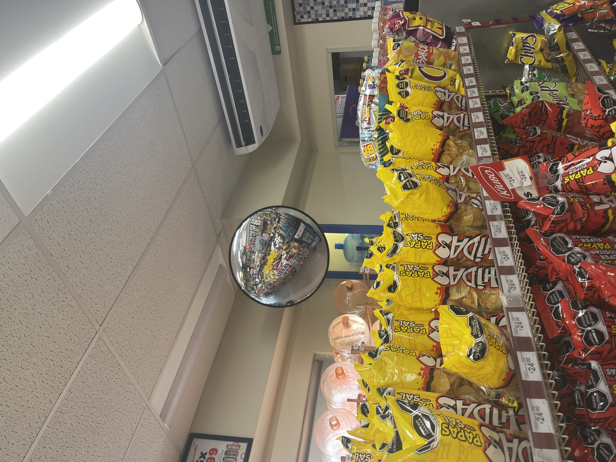 A low-angle shot inside a store, potentially a convenience store or small grocery in San Bernardino Tlaxcalancingo, Mexico, focuses on aisles of snacks. The right side of the frame features shelves stocked with chips, primarily brands such as "Papas," "Chidas," and "Chip's," with pricing tags of $24.00 visible. In the center, large water jugs are seen behind the chips. A round security mirror on the left reflects the store's interior. Overhead, a white ceiling with a grid pattern is visible, as well as a ventilation unit. A price tag is partially visible on the bottom left of the image. The chips are stacked on metal shelves, and the prices are written in black marker on white labels. The lighting is bright and harsh, likely from fluorescent lights.