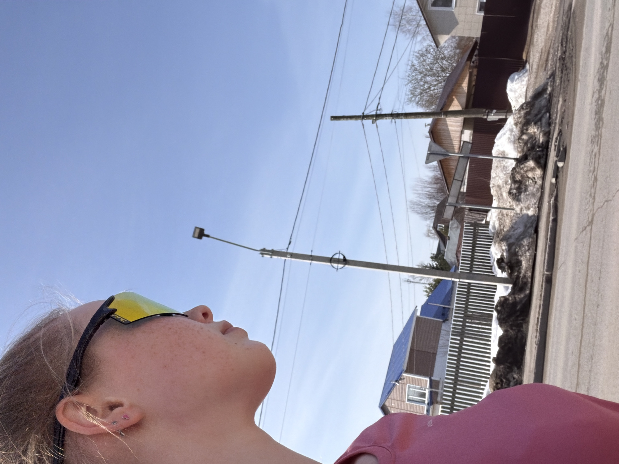 A medium-angle shot depicts a fair-skinned individual with freckles and yellow-tinted sunglasses, looking upwards. The person is wearing a pink top and has pierced ears. Above, a clear blue sky fills the frame, interspersed with telephone wires and utility poles. In the background, houses are visible, some with blue roofs, along with melting snow piles along the road. The location is Berezniki, Russia.