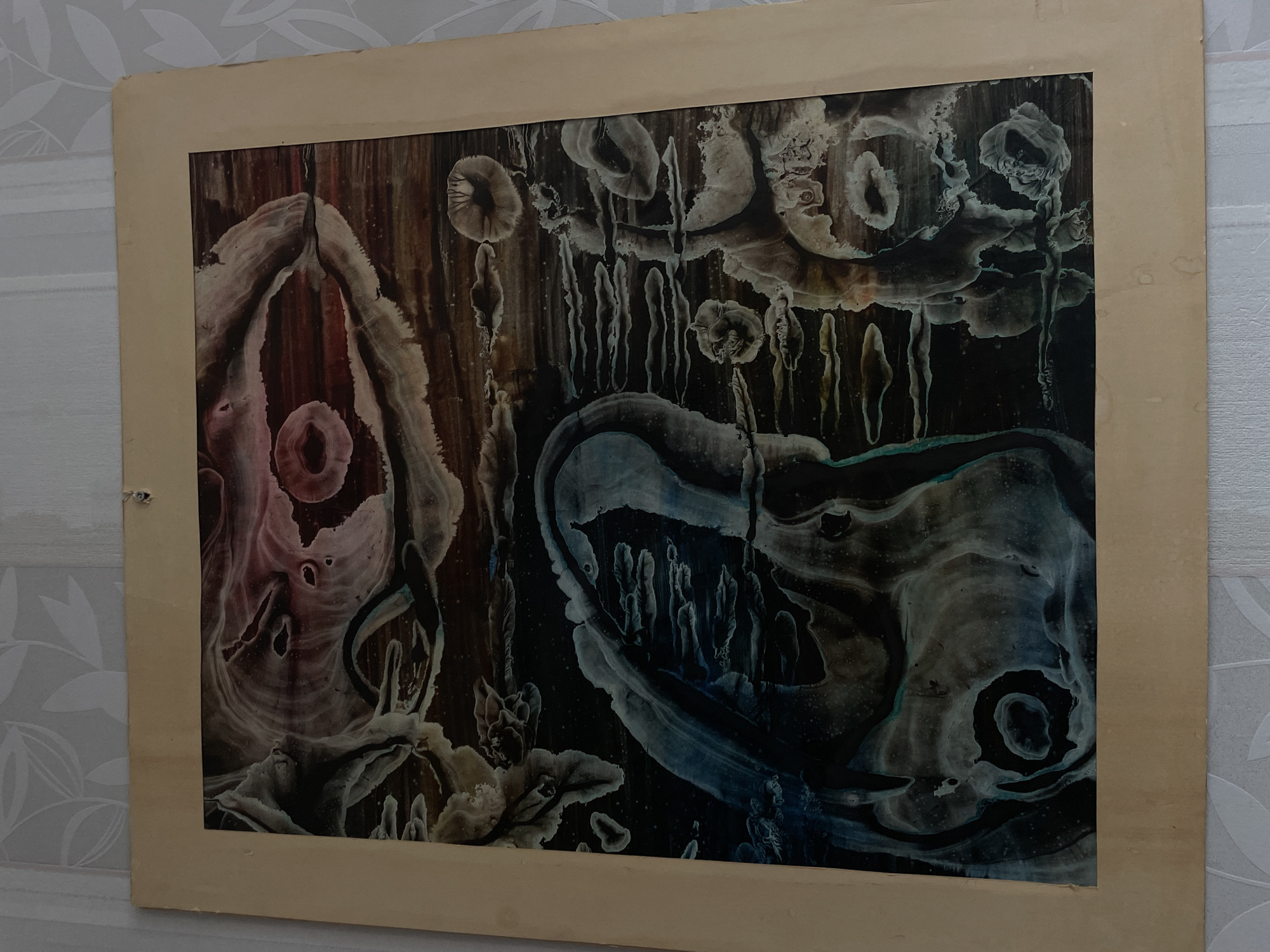 The image features a dark, abstract artwork mounted on a light brown board, set against a light-colored, patterned wallpaper. The artwork is the central subject, characterized by organic, fluid shapes and patterns primarily rendered in deep browns, blacks, and blues, highlighted with contrasting lighter shades of gray, white, subtle reddish-pink, and turquoise.
The abstract forms within the artwork evoke natural processes or structures, such as melting substances, clusters of fungi, cross-sections of wood or stone, or intricate geological formations. Many elements display circular or semi-circular contours, with prominent "drip" effects suggesting the movement of viscous liquids or natural erosion. On the left side of the artwork, reddish-pink and white shapes form a large, cavernous structure with a distinct circular element at its base. The upper and right sections contain numerous lighter, often mushroom-like or cellular structures with dark centers, from which vertical, stalactite-like streaks descend. The lower right portion of the artwork features large, fluid blue and gray forms, also containing internal circular patterns.
The artwork is presented on a plain, light beige or cardboard-colored mounting board. This board exhibits signs of age and wear, including slight discoloration, minor creasing, and a small tear or hole near the top-left corner. The edges of the mounting board appear somewhat worn.
The background of the photograph is a light-toned wallpaper adorned with a subtle, repeating pattern of leaves or floral motifs. There is no discernible activity or event taking place, and no text is visible within the image.