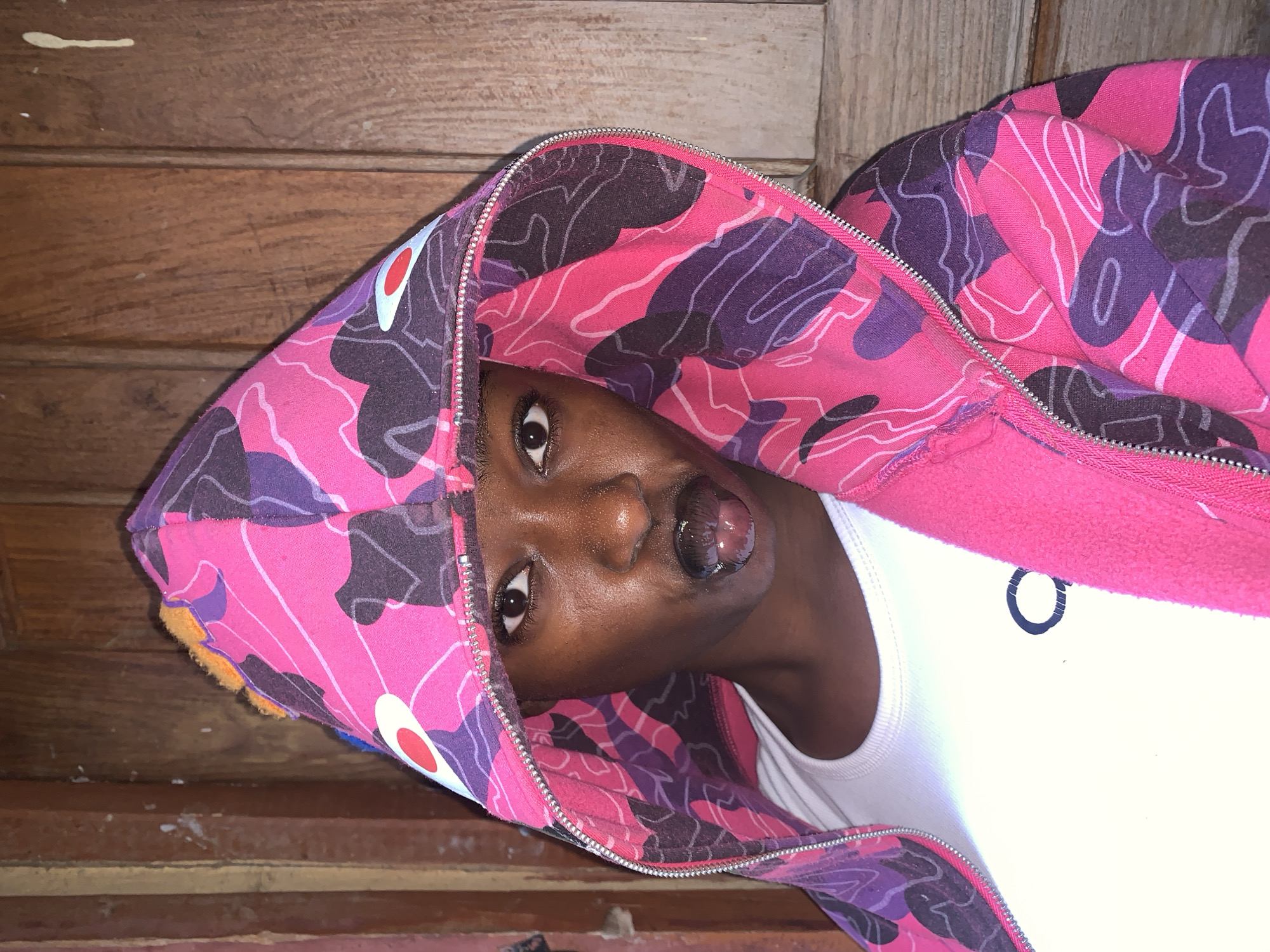 A person with dark skin is wearing a pink and purple camouflage hooded jacket, a white shirt, and sticking out their tongue. The person is indoors against a brown wooden surface. The location cannot be determined from the image and context.