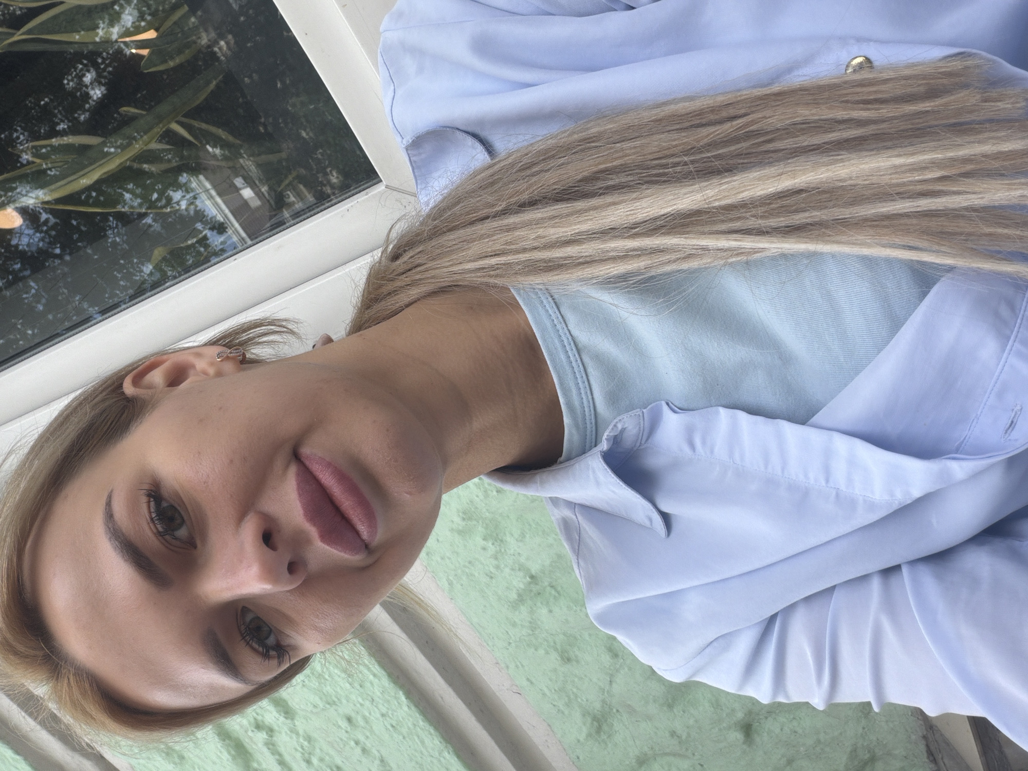 This is a close-up, rotated selfie of a young woman. She has long, blonde hair and is wearing a light blue shirt over a light blue top. Her makeup includes foundation, mascara, and a pinkish lipstick. Her expression is soft and perhaps a little playful, with her lips slightly parted. She is wearing a small, silver stud earring in her visible ear. The background includes a window with greenery and reflections of trees and sky, suggesting an outdoor setting or a room with a view. The light is natural and appears to be daylight, possibly morning or afternoon. The overall impression is a casual, candid moment captured by the subject herself. There is no visible text in the image.