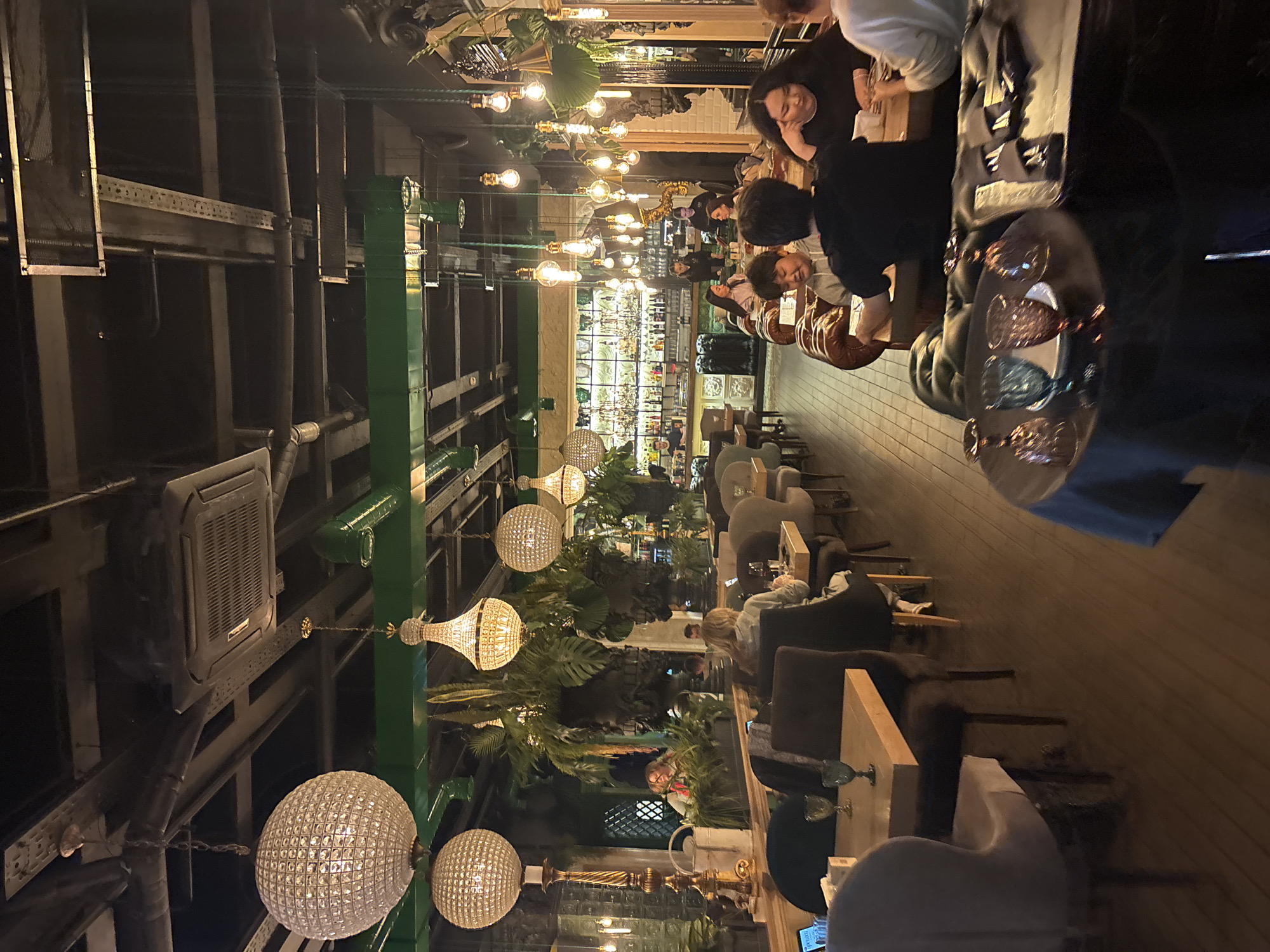 The image captures the interior of a restaurant located in Saint Petersburg, Russia. The space is adorned with decorative chandeliers and a dense array of plants, giving it a vibrant, botanical feel. Patrons are seated at tables, engaging in conversation and dining. The restaurant has a full bar in the background, stocked with bottles and glassware. Industrial style air conditioning and support structures are visible on the ceiling.