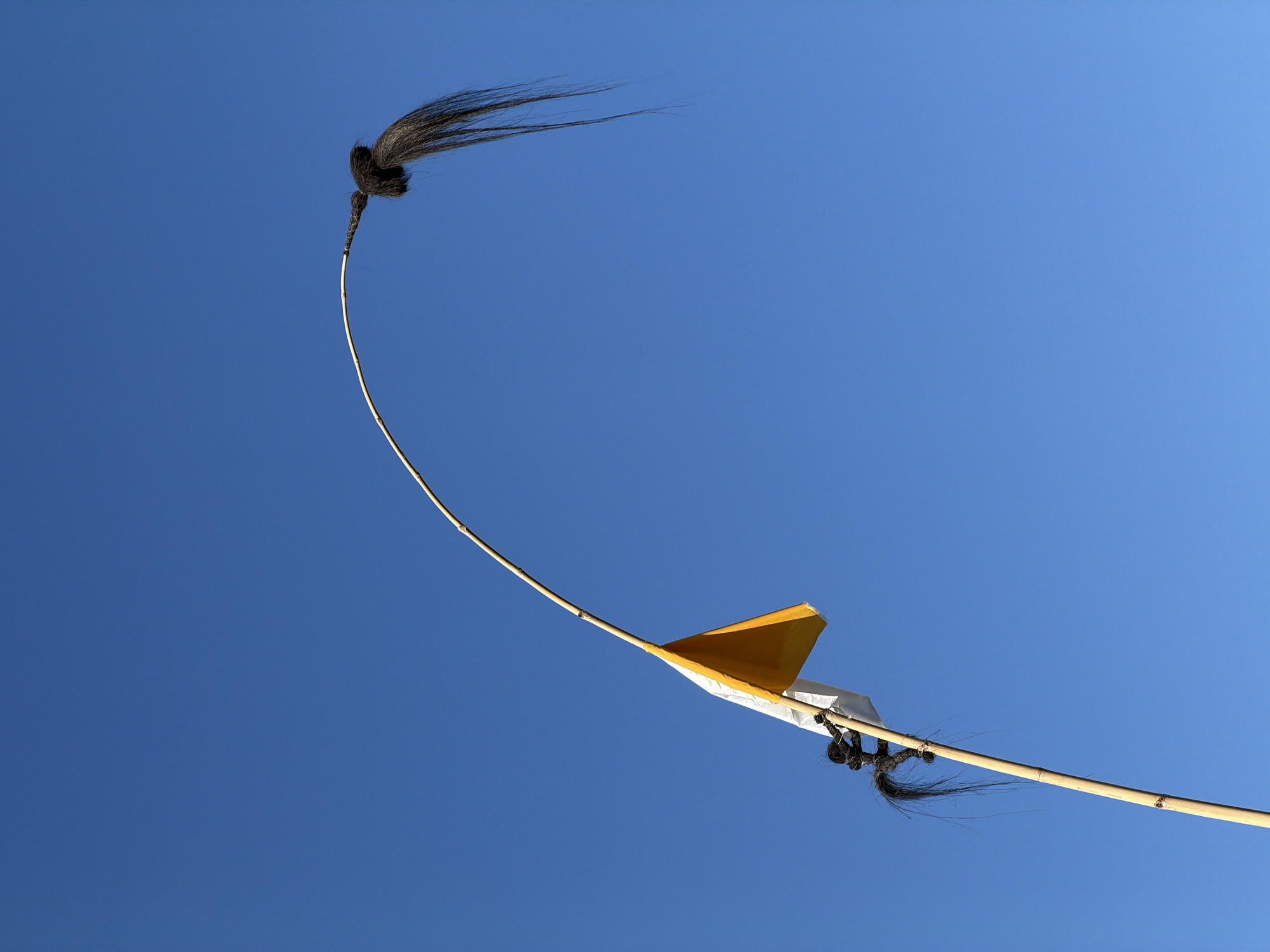 A low-angle shot shows a penjor against a clear, bright blue sky. The penjor, a tall, curved bamboo pole decoration used in Balinese Hindu ceremonies, is topped with a tuft of black, fibrous material resembling hair. A yellow and white banner is attached to the middle of the pole. Another, smaller tuft of the same black material is tied further down the pole, close to the bottom of the visible section. The pole appears to be made of bamboo, with visible nodes along its length. The photograph was taken in Mengwi, Indonesia.