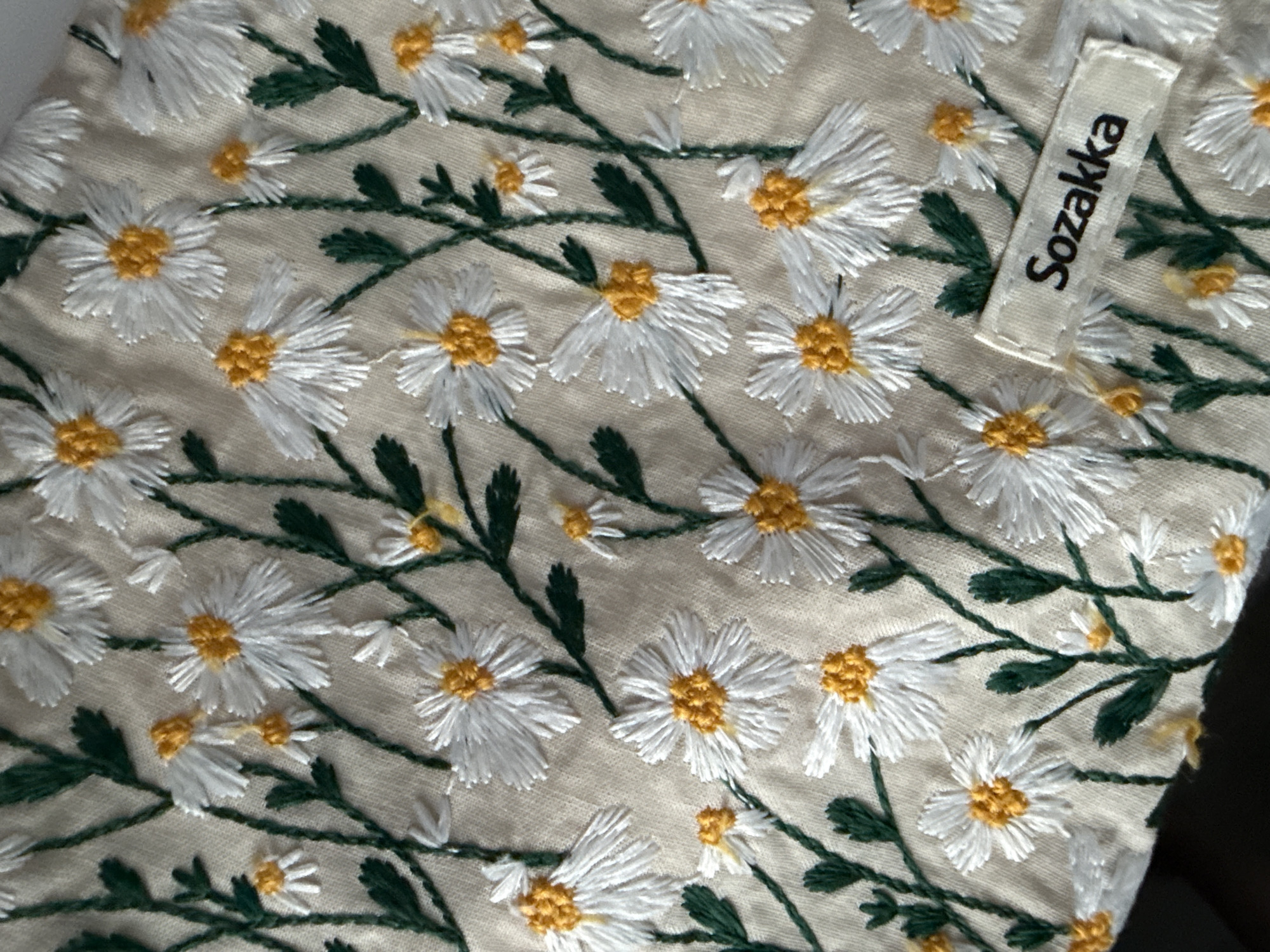 The image shows a close-up view of a beige fabric embroidered with a pattern of white and yellow daisies and green vines and leaves. A small, rectangular tag with the text "Sozakka" in black font is sewn onto the fabric. The scene does not show any evidence of being located in Moscow, Russia.