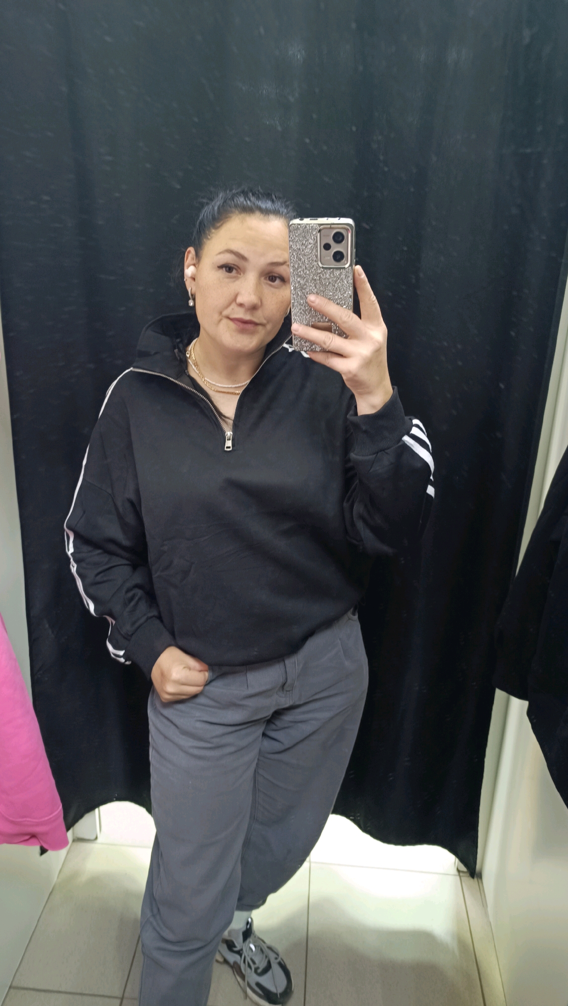 A woman is taking a mirror selfie in what appears to be a fitting room. She has her dark hair pulled back in a bun and is wearing a black, half-zip sweatshirt with white stripes on the sleeves. She is also wearing gray pants and athletic shoes. The fitting room has a black curtain behind her and white walls. A pink garment is visible to her left. The image appears to be taken in the city of Chaikovsky, Russia.