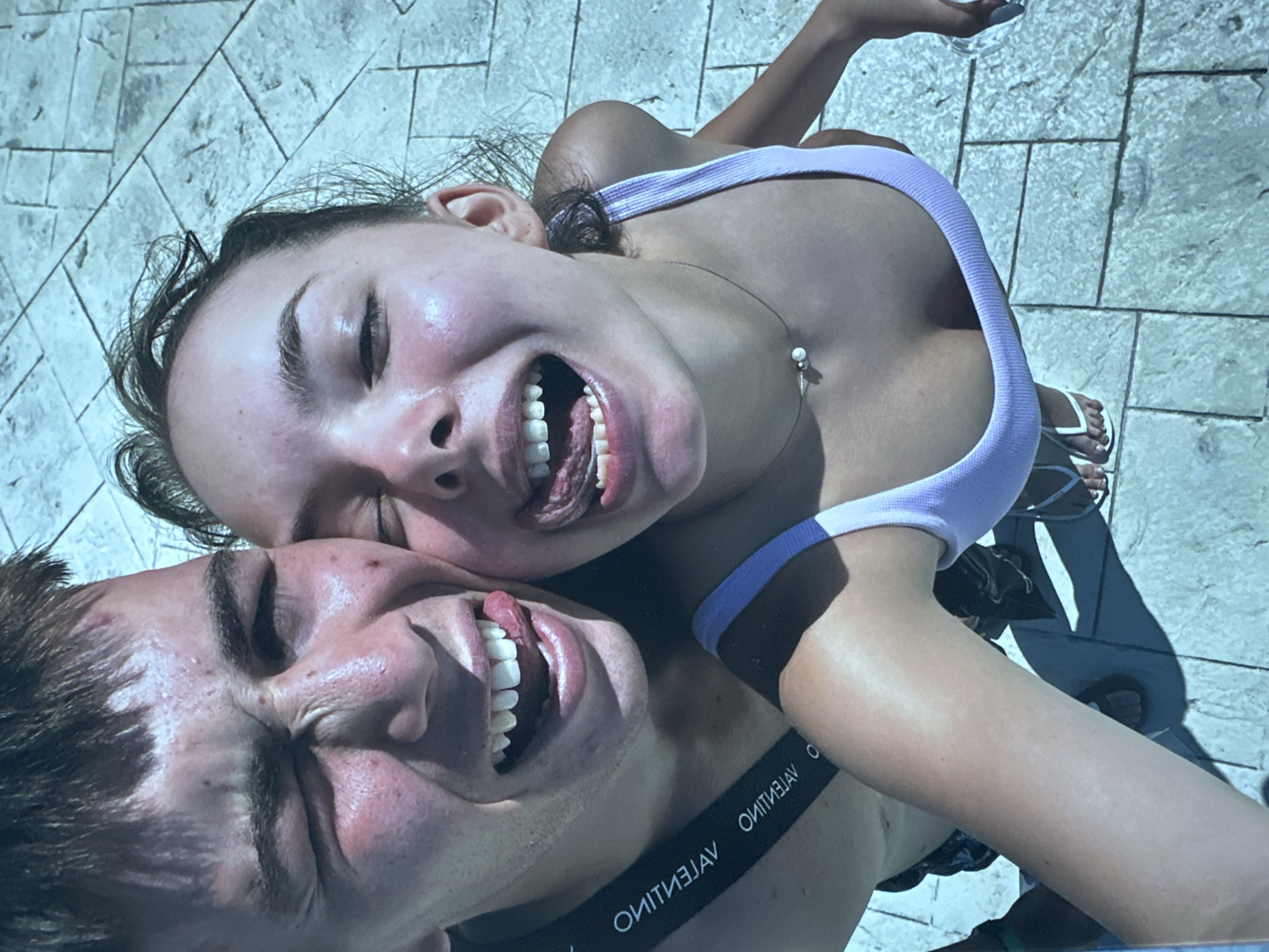 The image shows a close-up, high-angle selfie of two young adults, a male and a female, outdoors. They appear to be by a pool, as evidenced by the tiled ground and the female's swimwear. Both individuals are sticking out their tongues and making exaggerated facial expressions. The male is on the left, with his eyes squeezed shut and tongue sticking out. He is wearing a black strap with white lettering. The female is on the right, also with eyes closed and tongue out, wearing a light purple bikini top and a pearl necklace. The lighting suggests it is a sunny day. There is no visible indication of the city of Sofia, Bulgaria, or any specific location within it.