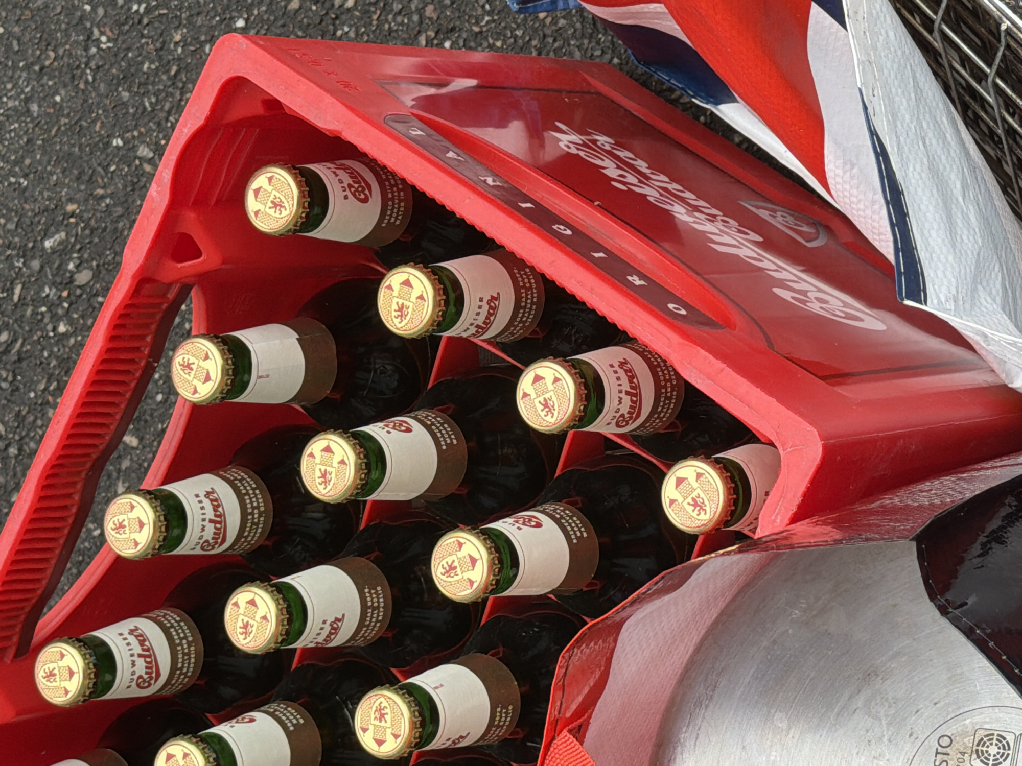 A high-angle, close-up shot shows a red plastic crate filled with several bottles of Budweiser Budvar beer. The crate, marked with "Budweiser Budvar ORIGINAL", is partially covered by a white and red cloth. The bottles are green and brown, with white labels, gold caps, and the Czech coat-of-arms. The crate is sitting on asphalt. The location is not specified.