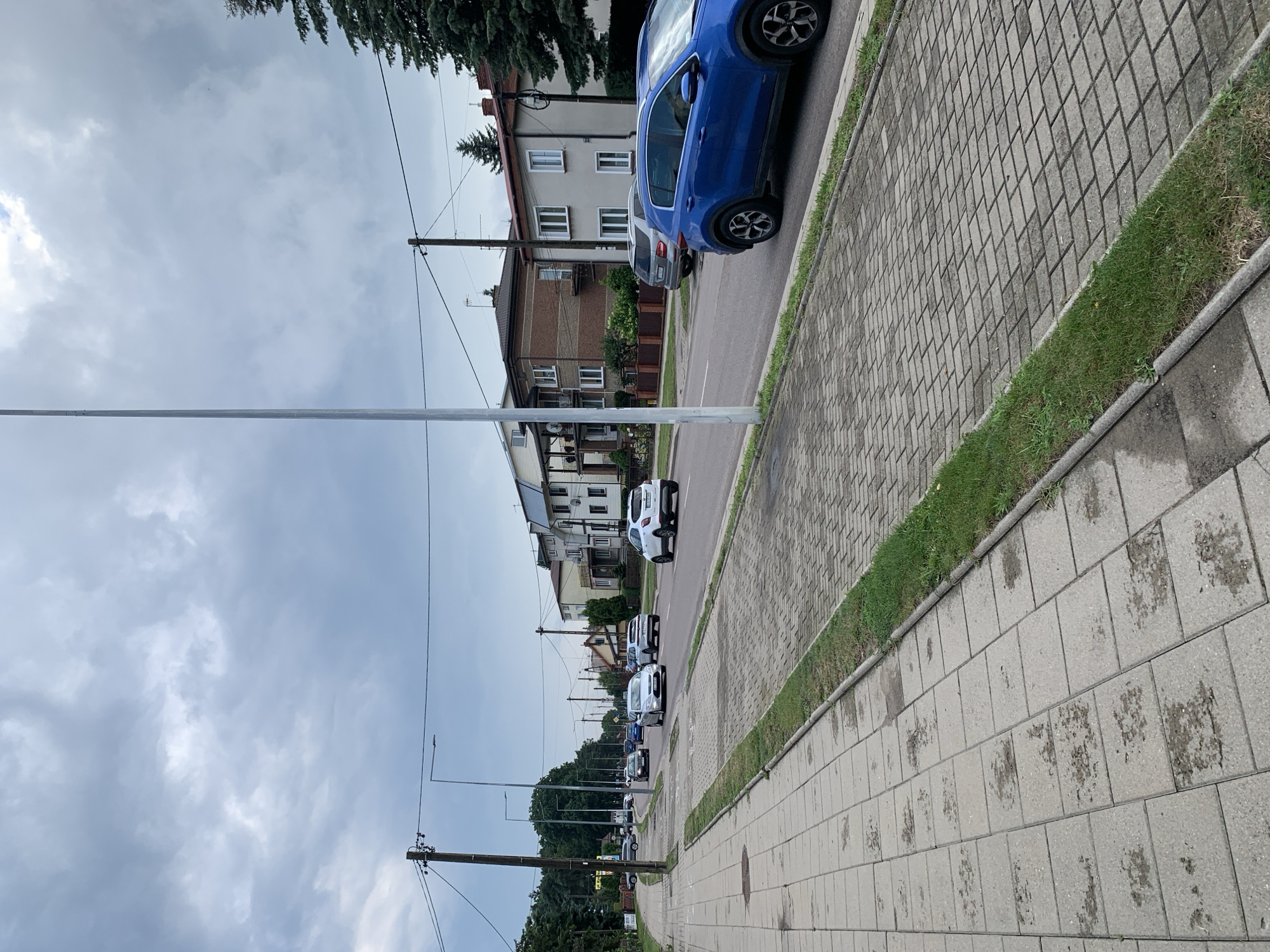 The image depicts a street scene in Białystok, Poland, on a cloudy day. The perspective is from a sidewalk, looking down a road lined with trees and residential buildings. Multiple cars are visible on the road, some in motion and others parked. The sidewalk is paved with concrete tiles, and a paved bike path runs alongside it, bordered by grass. Streetlights and utility poles are present along the road. The sky is overcast, with heavy clouds suggesting a possibility of rain. The overall atmosphere is that of a typical urban environment on a temperate day.