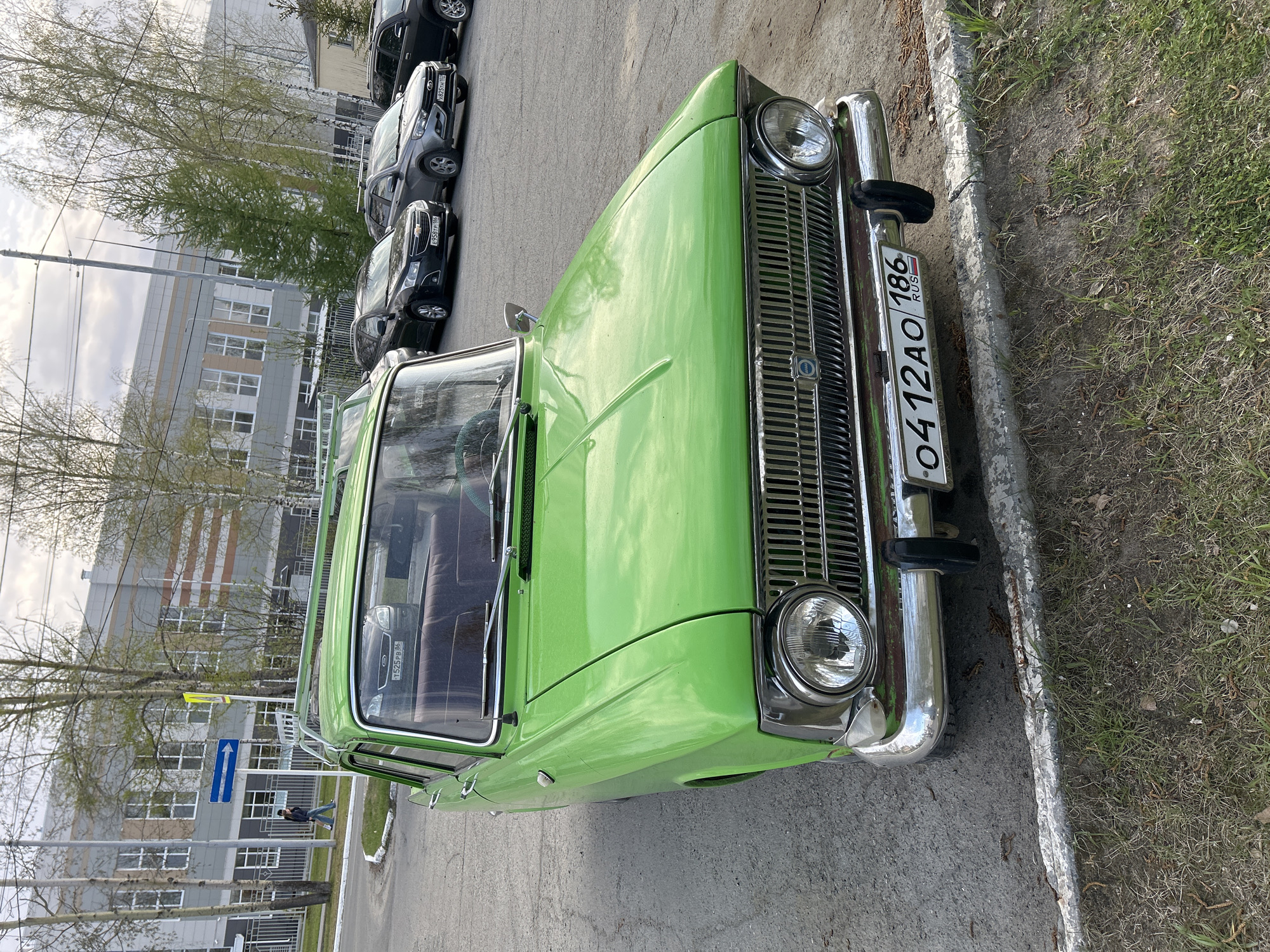 A green vintage car is parked on the street. The license plate number is 0412AO 186, indicating registration in Russia. Several other cars of modern make are parked behind it. A multi-story building is present in the background, and a directional sign can be seen on the left side of the image. The location of the image is in the city of Uray, Russia, as indicated by the license plate.