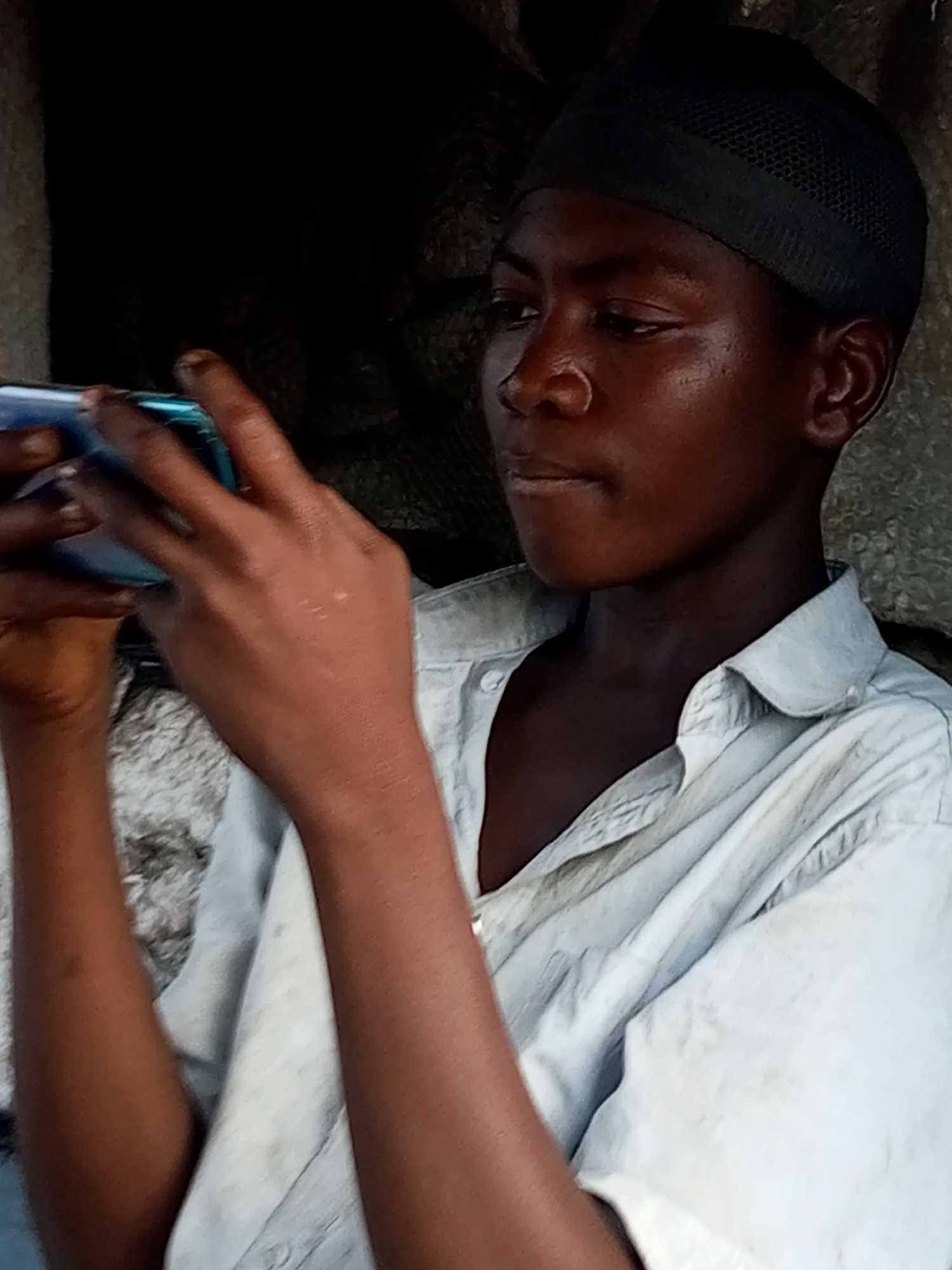 The image shows a close-up, medium shot of a young person wearing a light-colored, button-down shirt and a black skullcap. The person is holding a smartphone horizontally with both hands, appearing to be looking at the screen. Their face is turned slightly to the right, with their gaze directed downwards. The lighting suggests it might be daytime, possibly indoors or in a shaded outdoor area, given the contrast between the bright skin and the darker background. The setting appears somewhat rustic or simple, with a textured, possibly rough wall or material visible in the background. The person's expression is neutral, focused on the device. The image was taken in Gombe, Nigeria.
